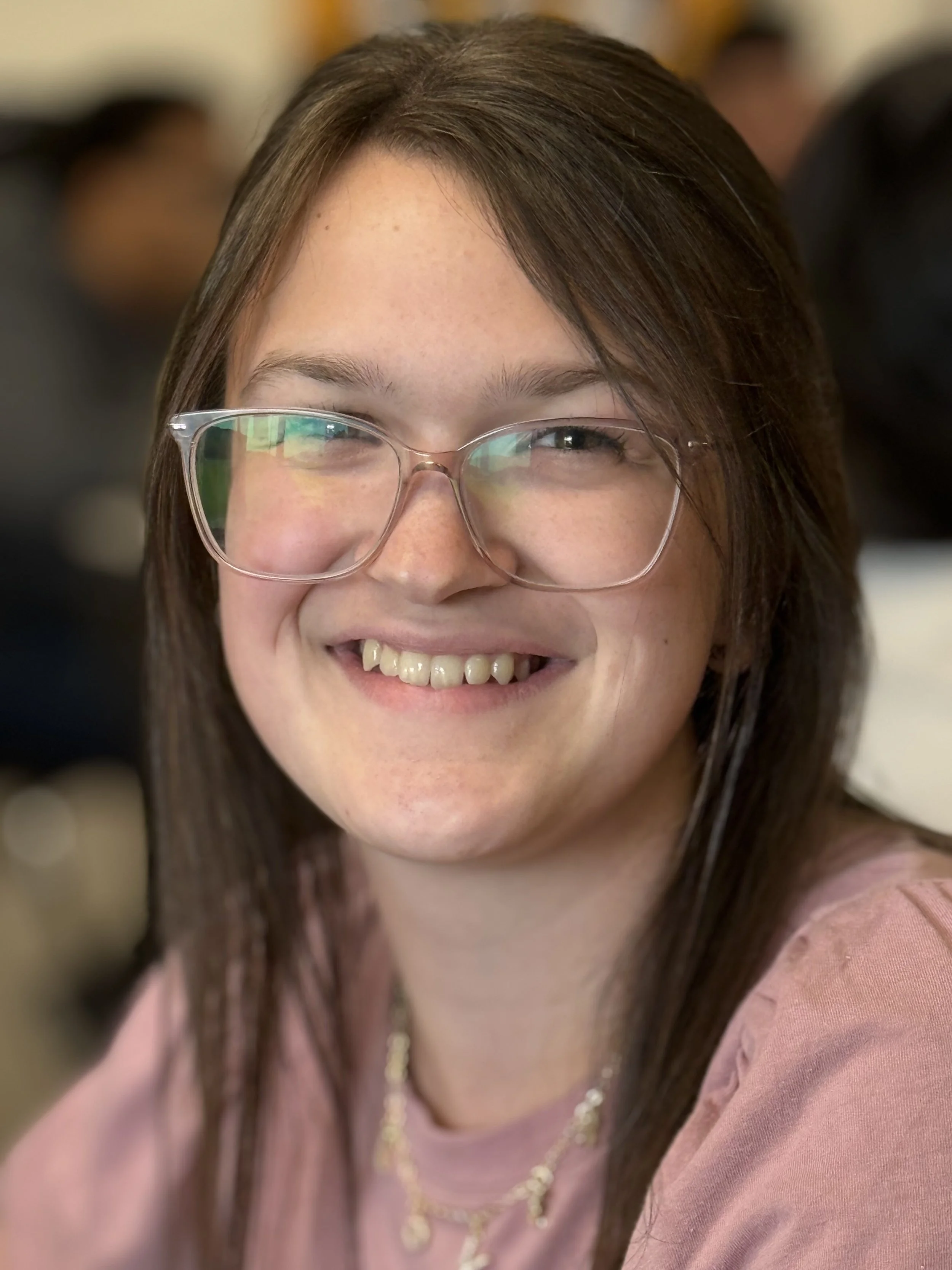 Close-up of a young woman smiling, wearing glasses and a pink top, with a blurred background.