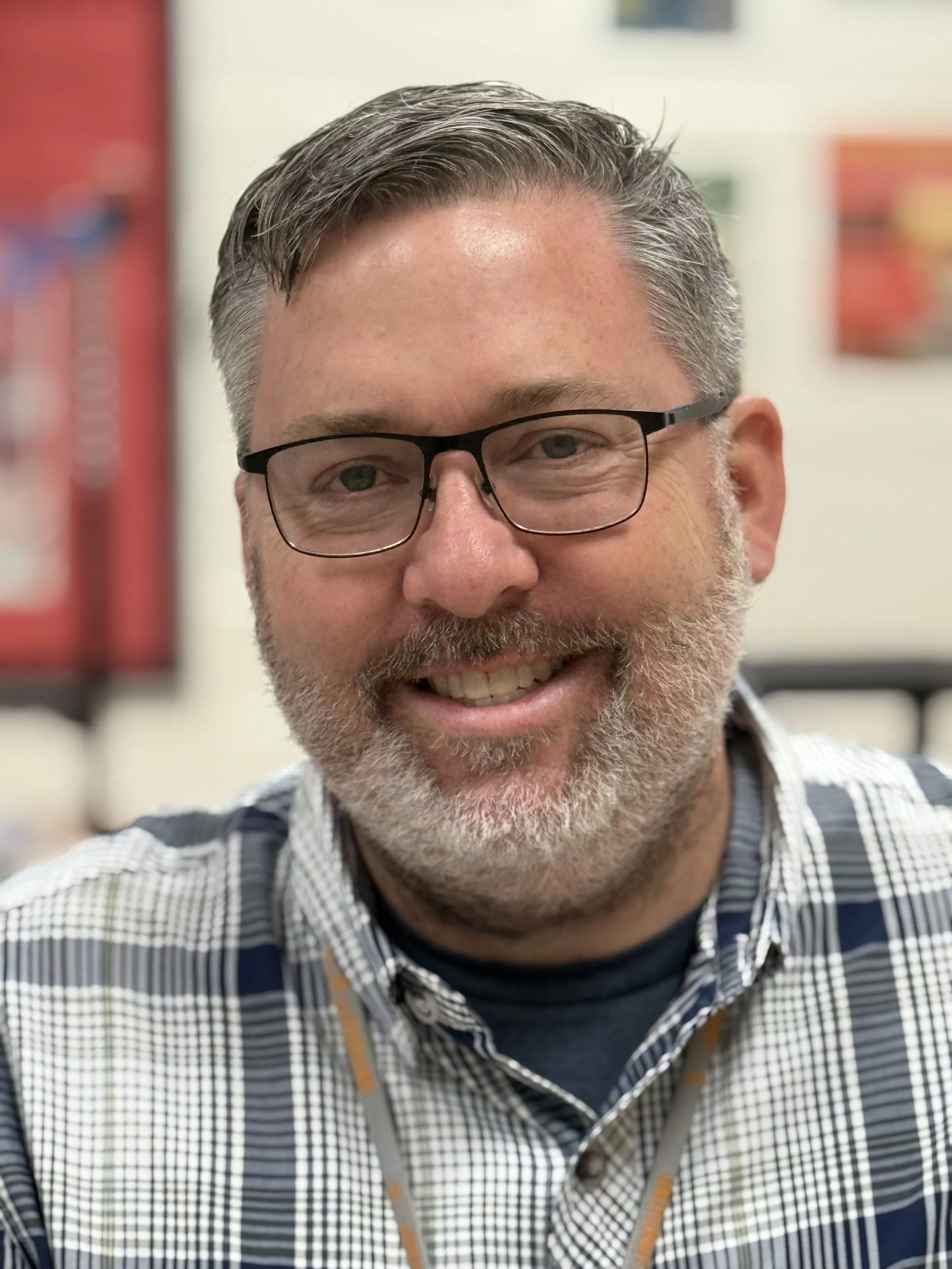 A smiling middle-aged man with glasses and gray hair, wearing a plaid shirt, in an indoor setting.