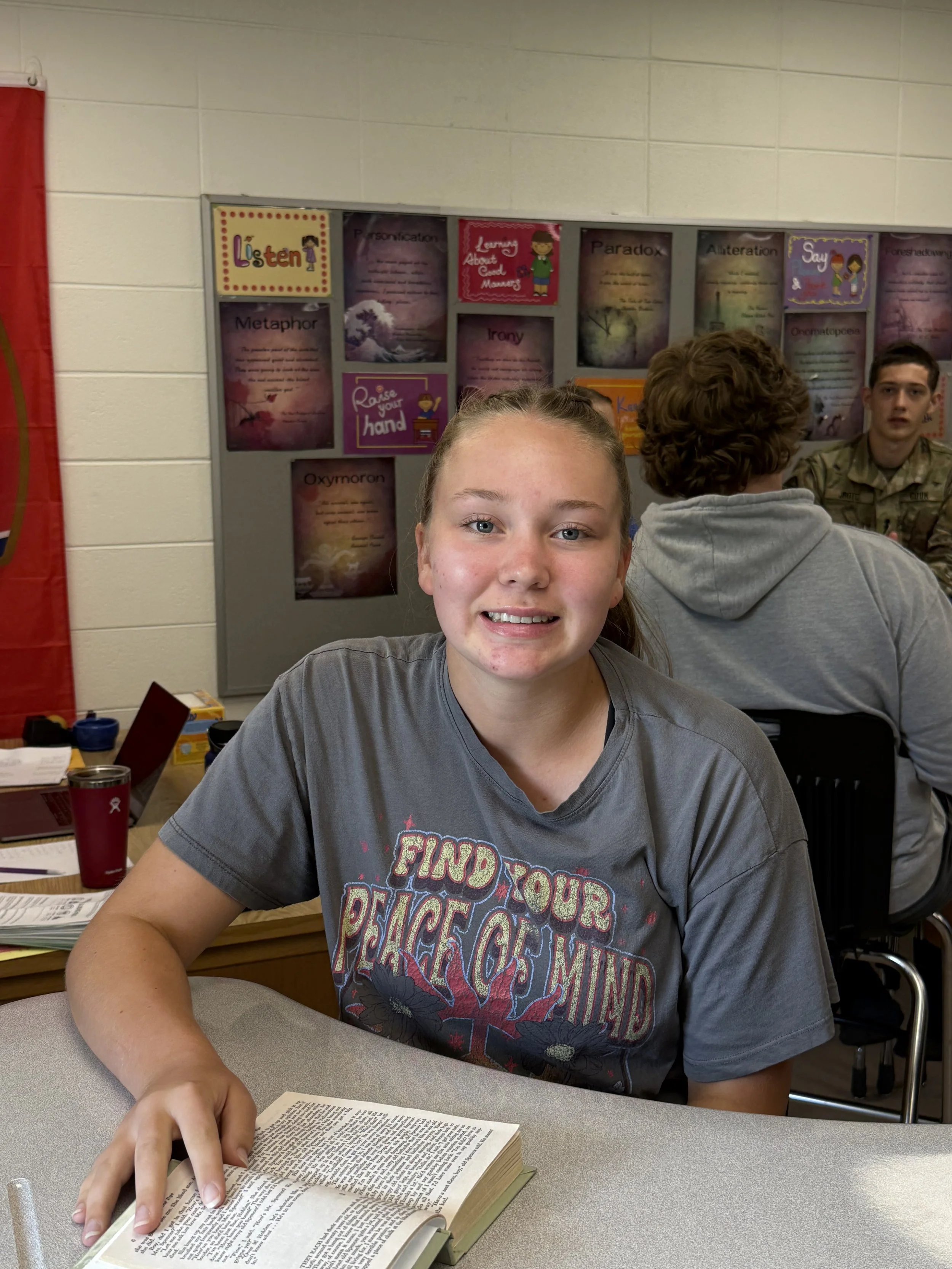 A teenage girl with braided hair sitting at a classroom desk, using a laptop, with other students in the background.