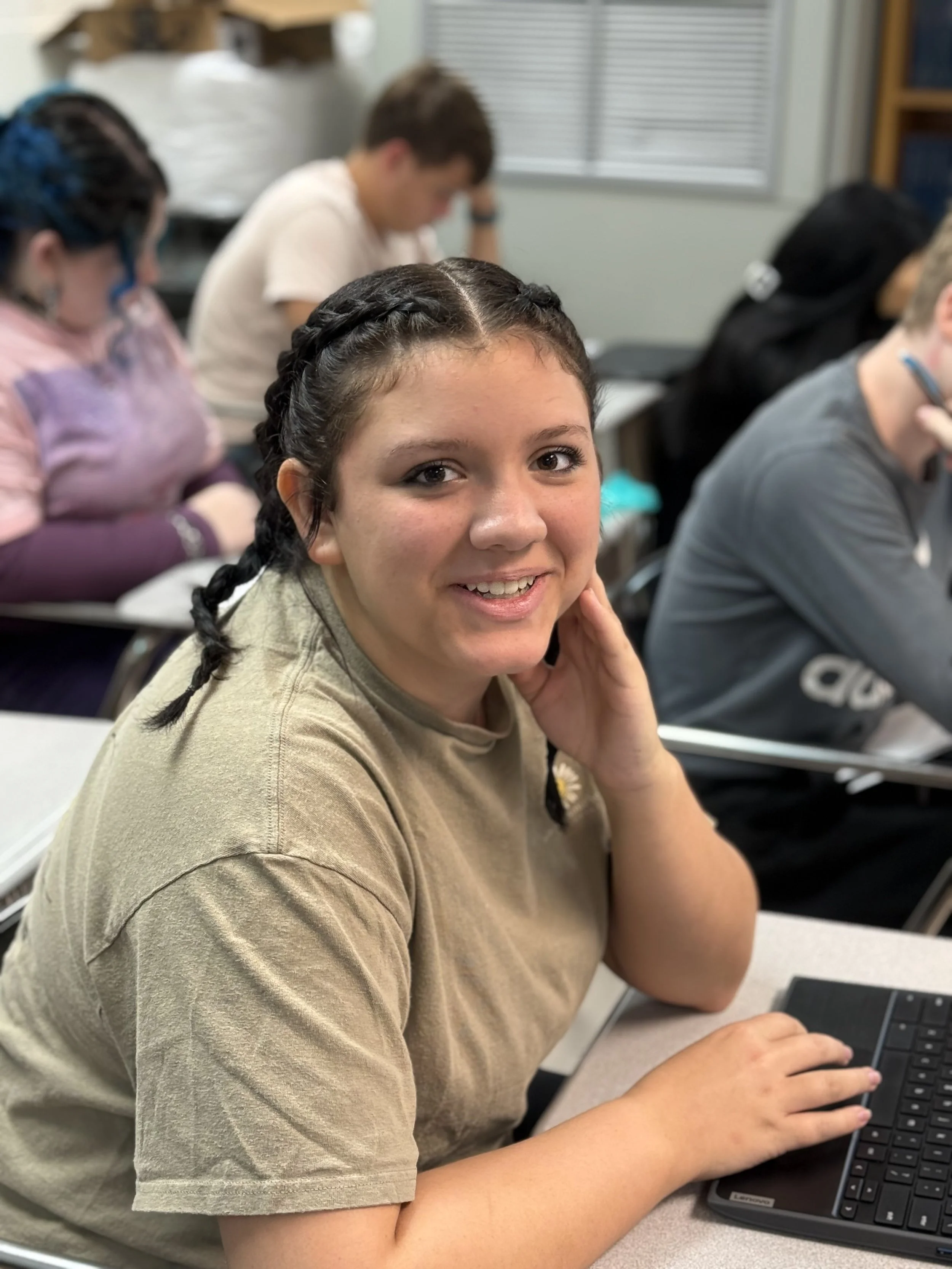 A girl with braided hair and a beige T-shirt smiling at the camera in a classroom with other students working at their desks.
