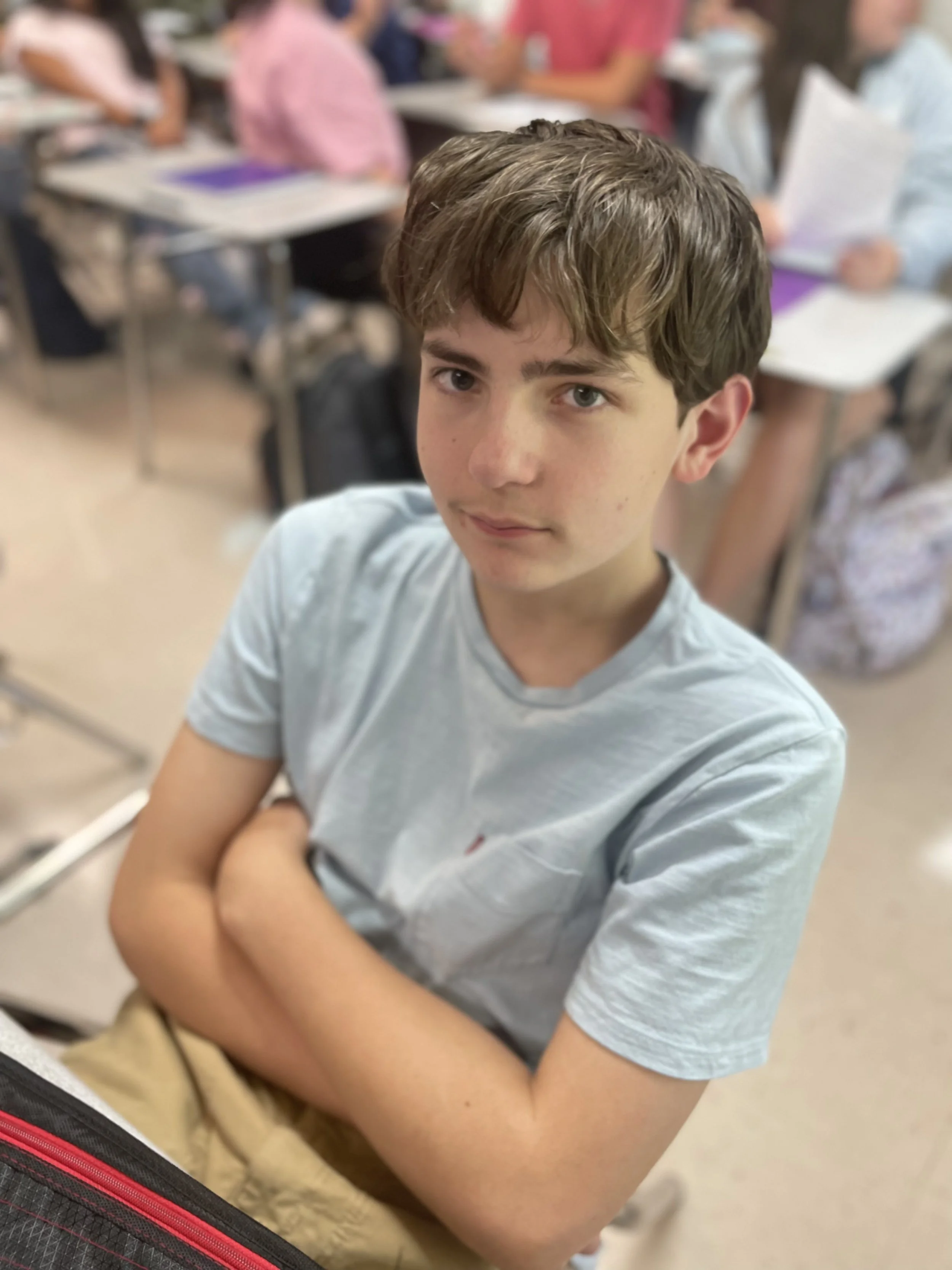 A young boy with short brown hair sitting in a classroom with his arms crossed, surrounded by students at desks.
