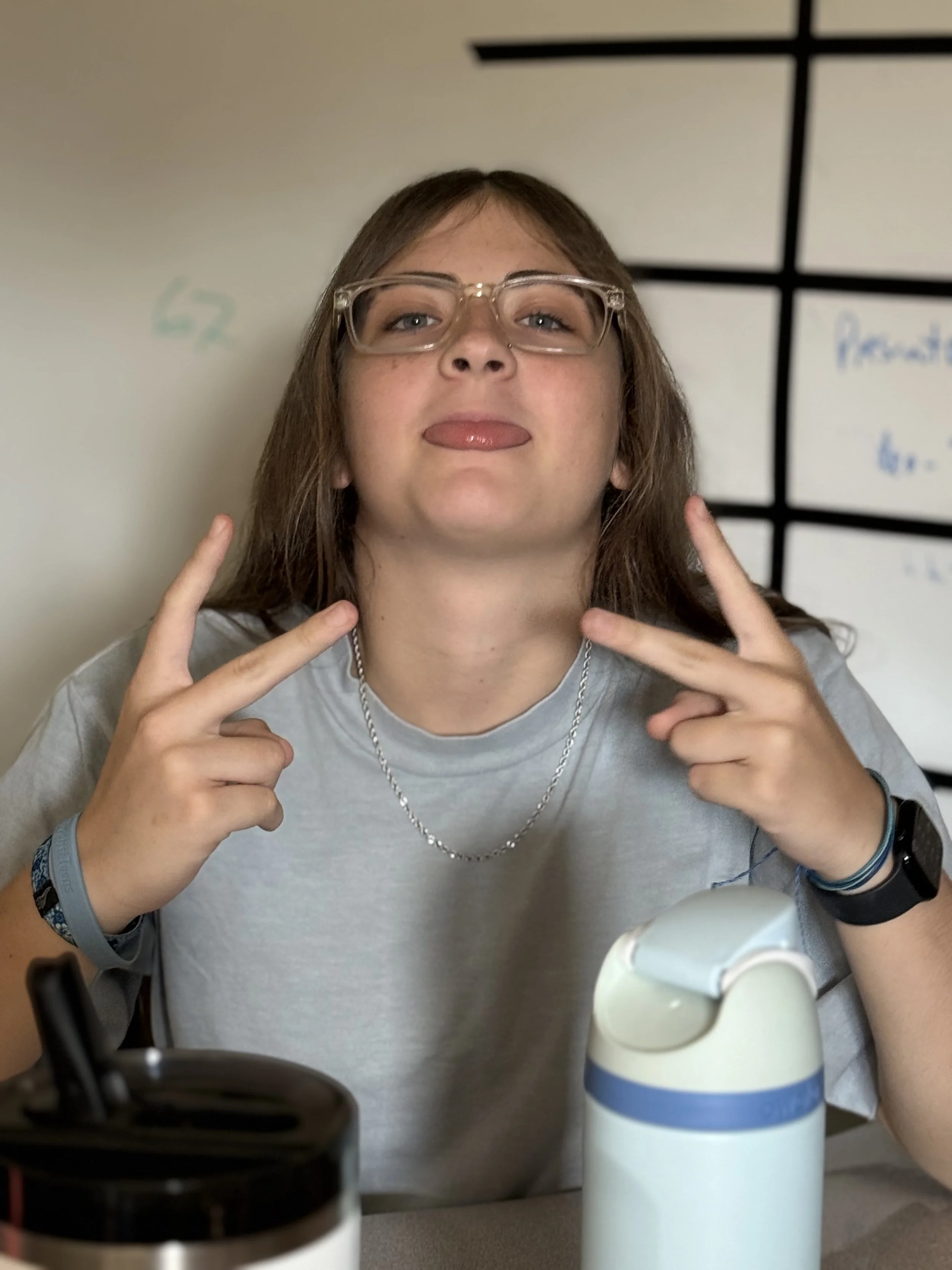 Young woman with glasses and brown hair making a peace sign near her face, sitting at a desk with water bottles in front, against a whiteboard background.