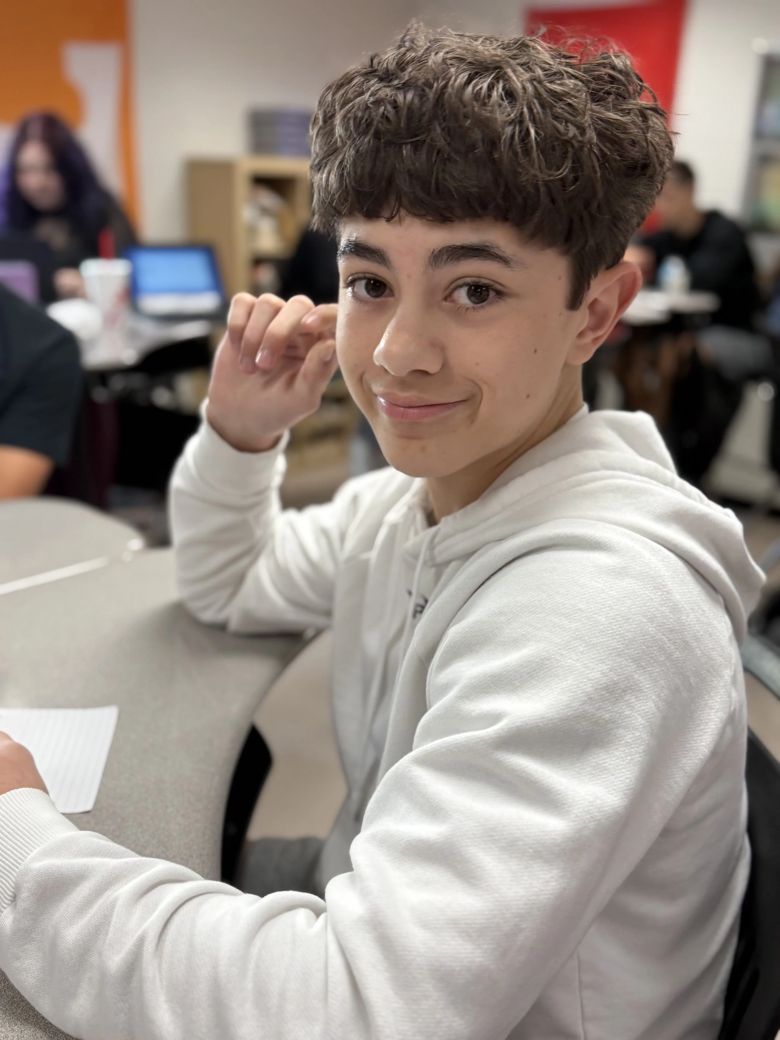 A teenage boy with brown, curly hair smiling while sitting at a table in a classroom.