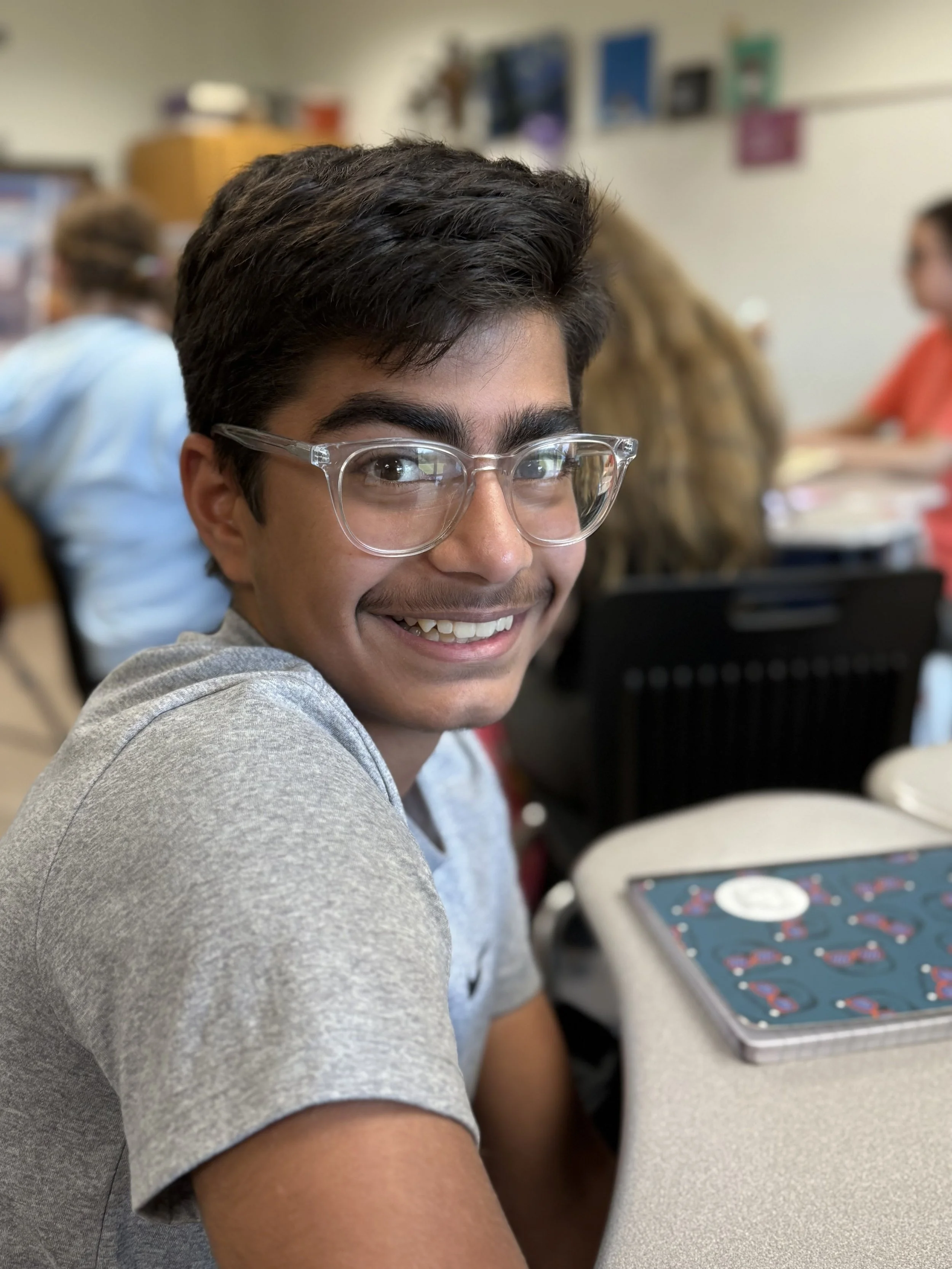 A young man with glasses smiling, sitting at a desk in a classroom with other students in the background.