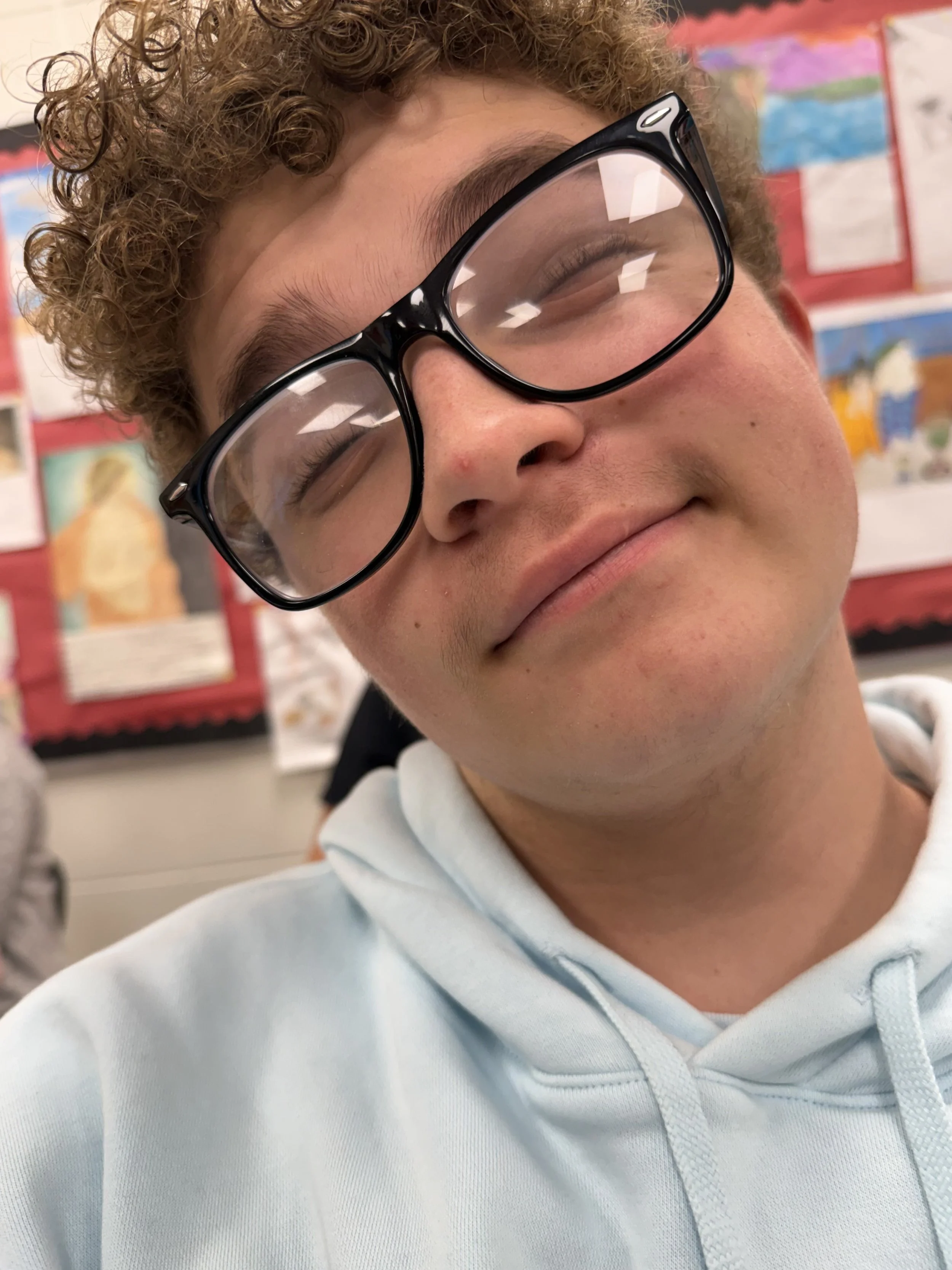 A young person with curly hair and glasses resting their chin on their hand, looking at the camera.