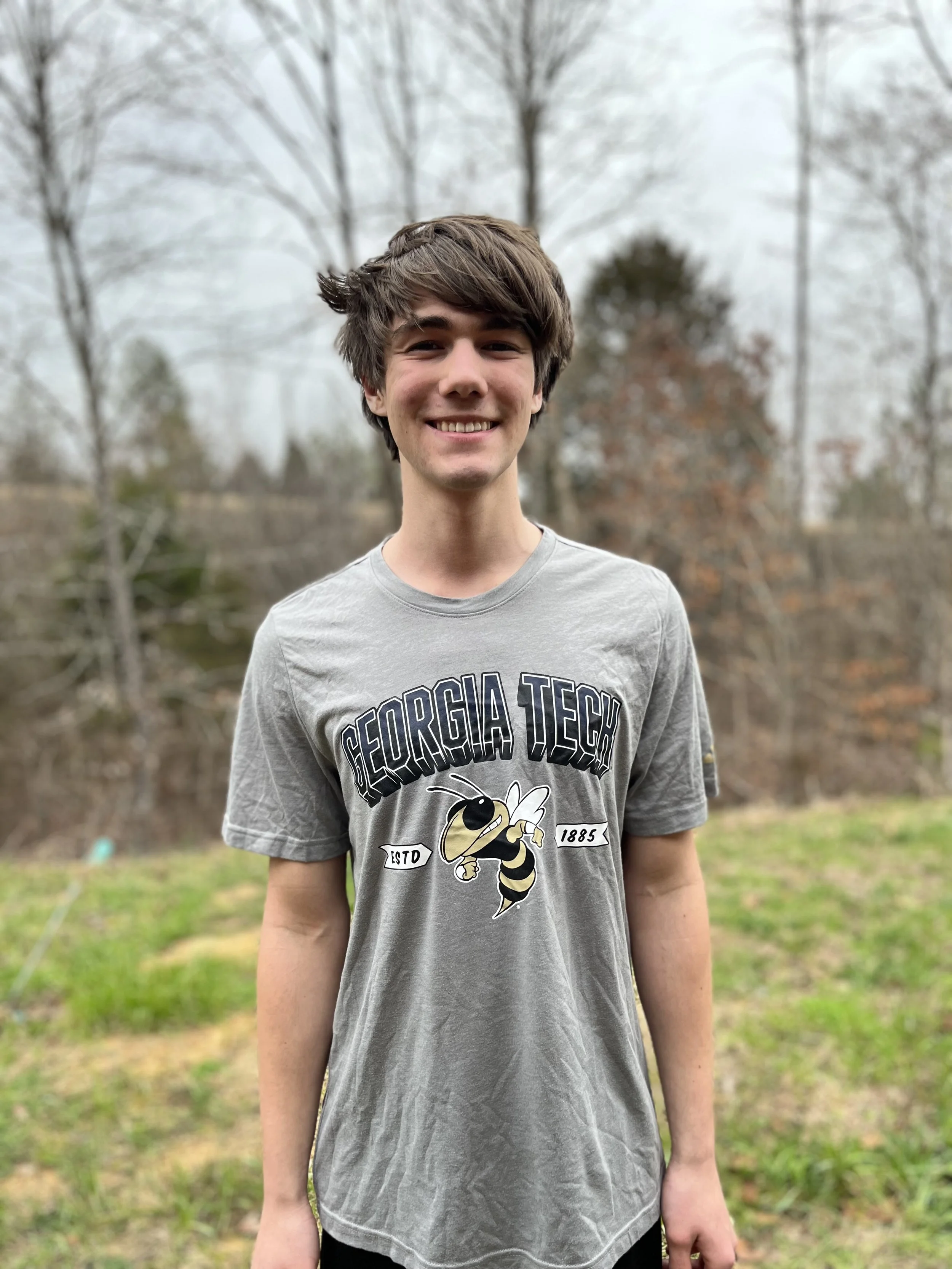 A young man with brown hair and a light beard smiling while looking over his shoulder in a classroom or office setting.