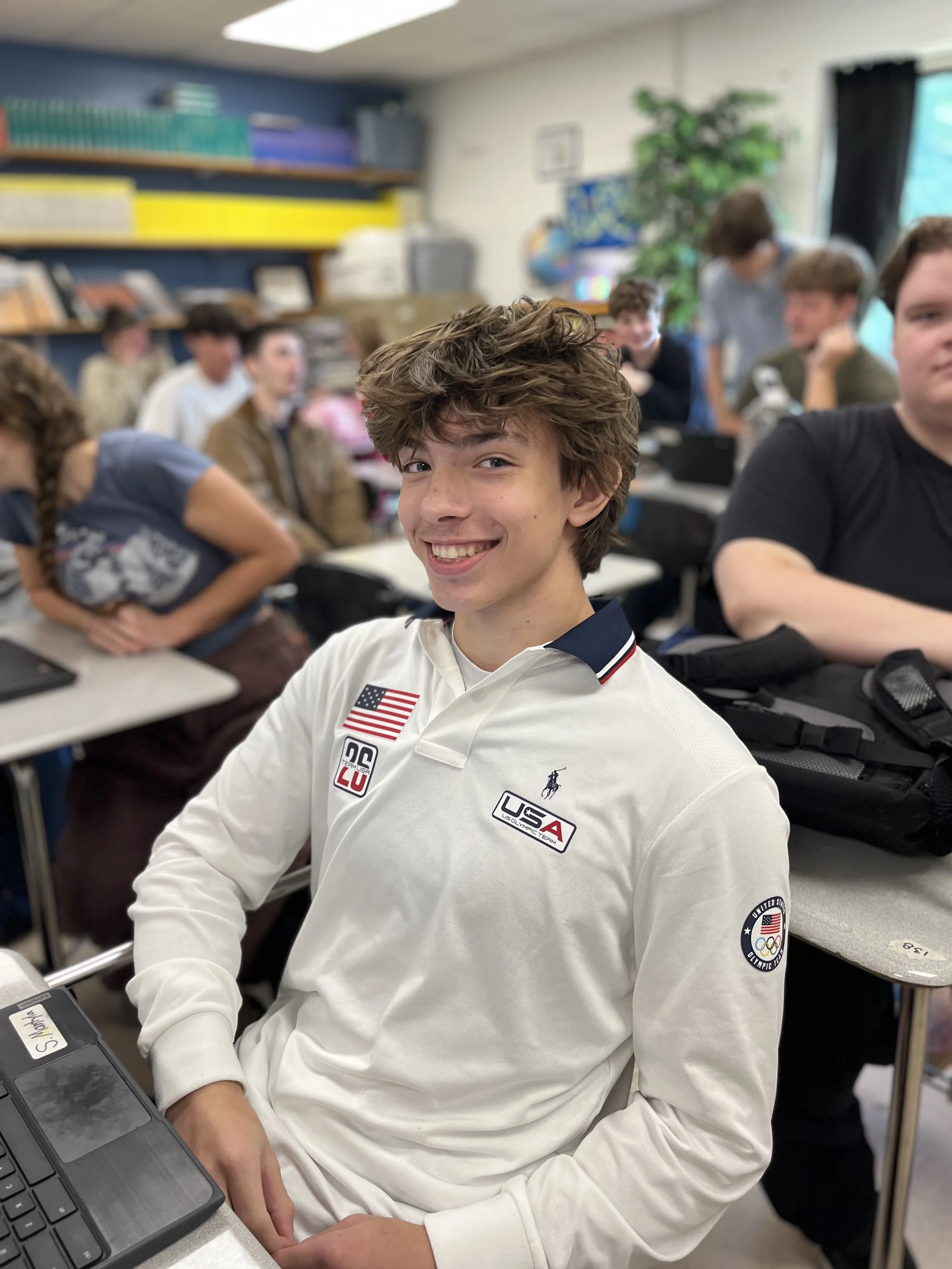 A teenage boy with short brown hair smiling and making a peace sign with his right hand in a classroom.