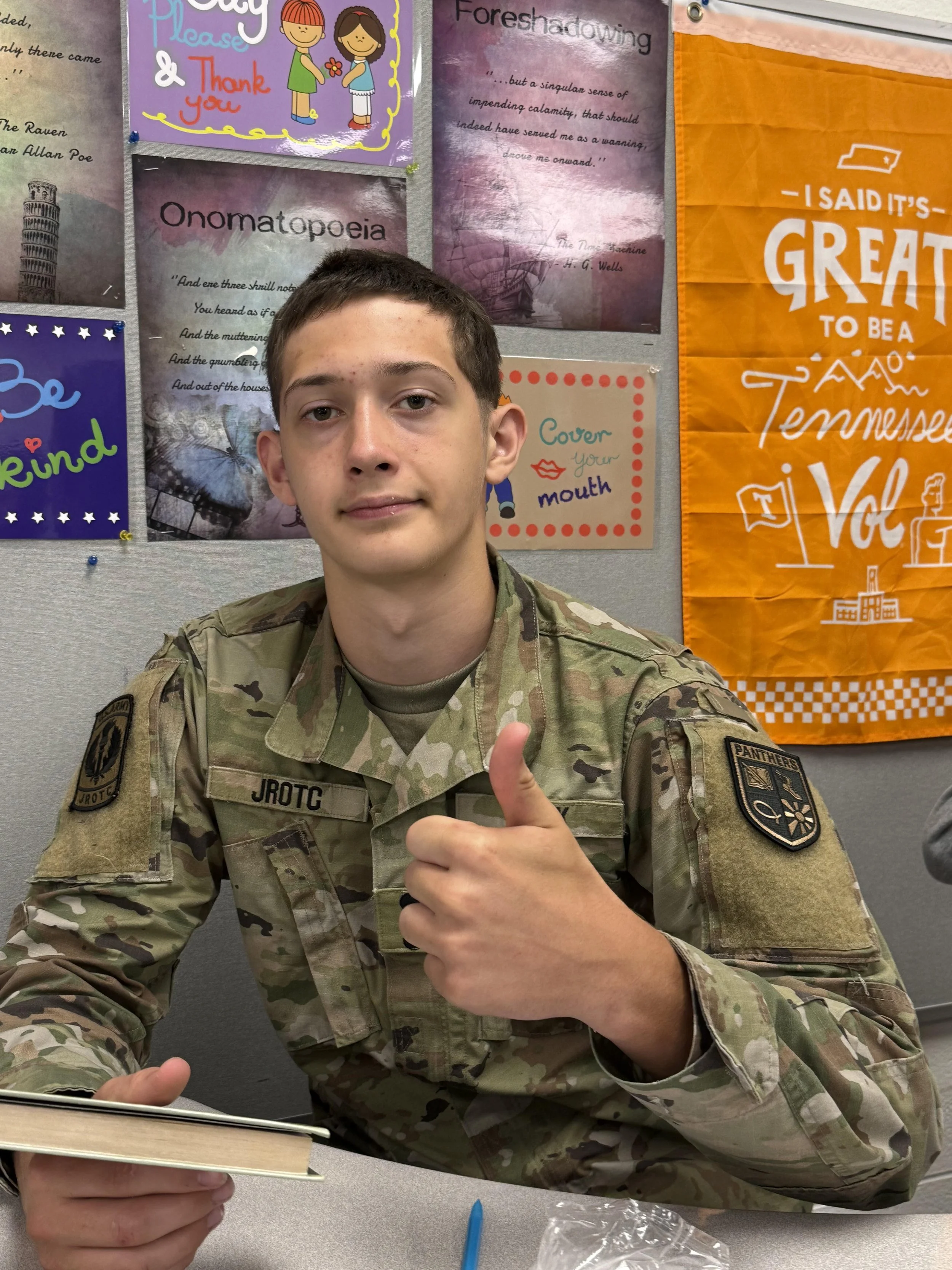 A young man with short brown hair giving a thumbs up while sitting at a table in a room with a red flag and military posters on the wall.