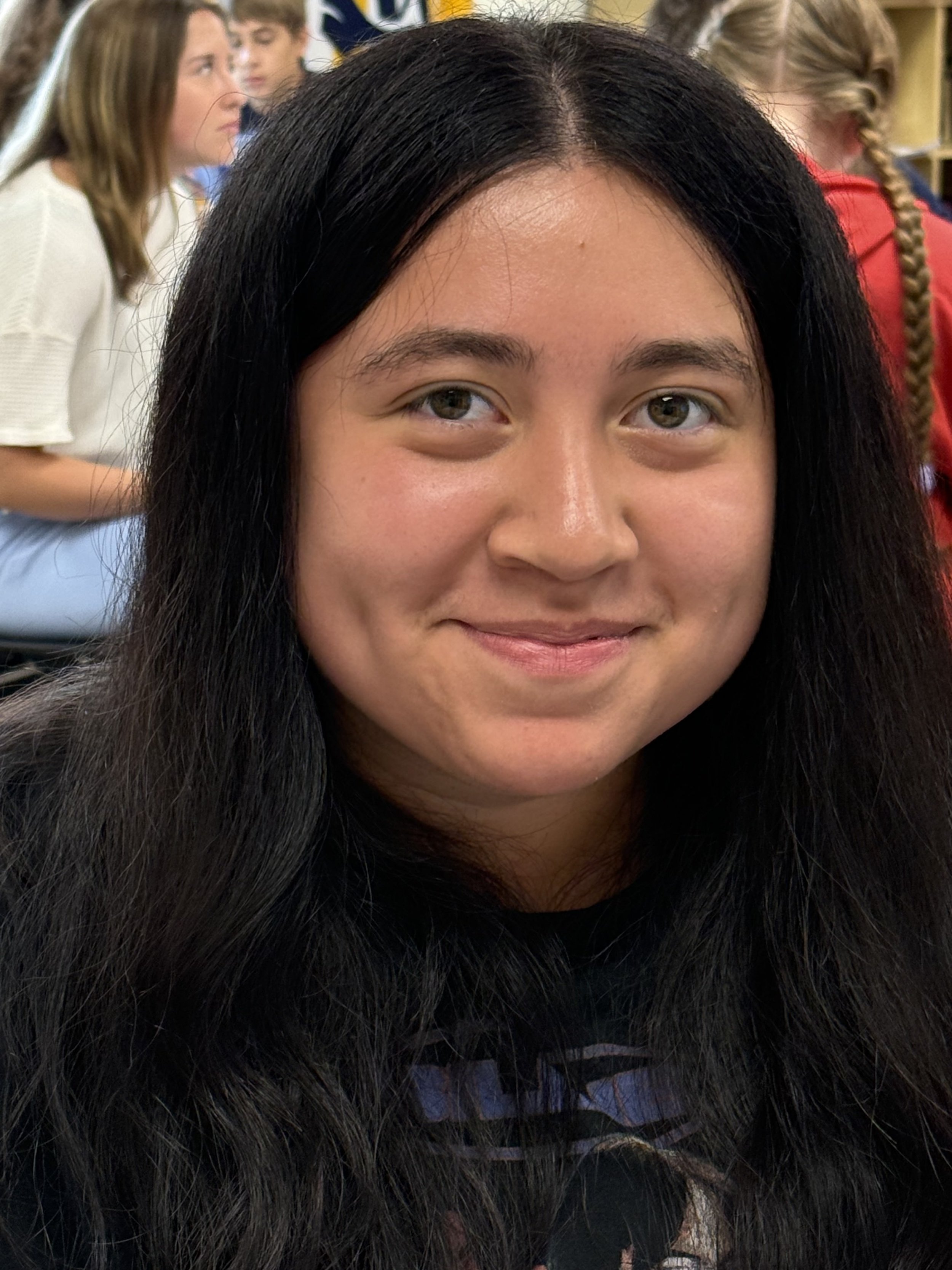 A young woman with long dark hair and a turquoise shirt sitting at a desk with a notebook and colorful pens, in a classroom with other students in the background.