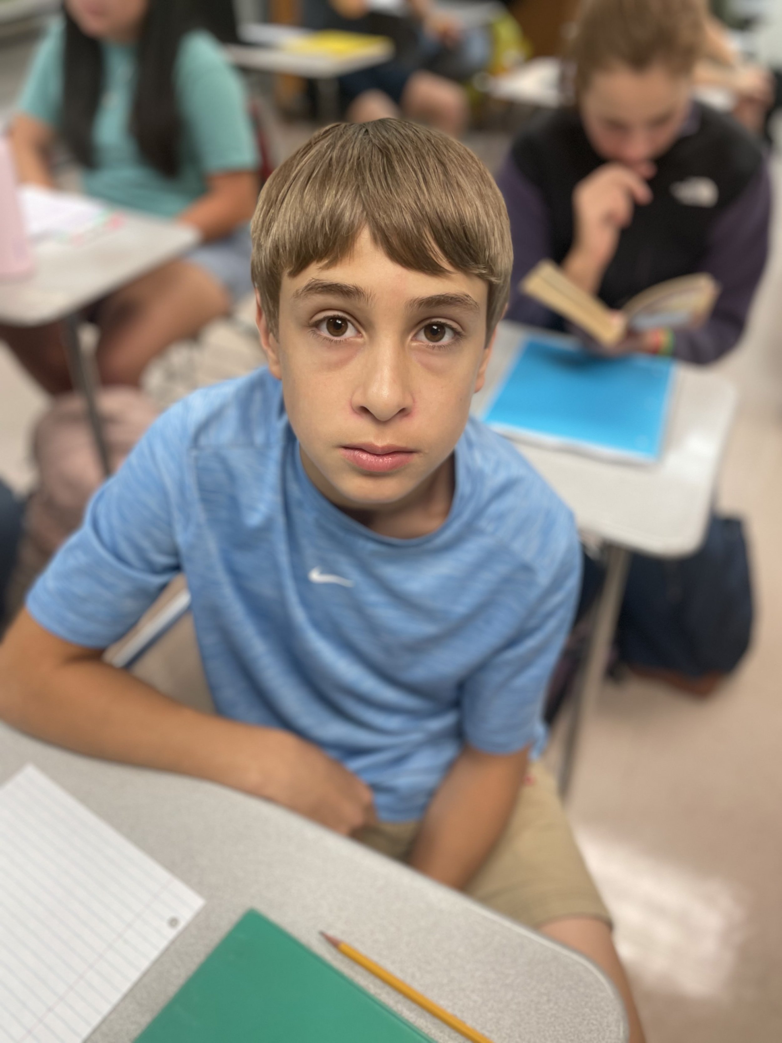 A young boy with brown hair and wearing a blue shirt looks up at the camera in a classroom, sitting at a desk with a green notebook and a pencil in front of him. Other students are visible in the background.