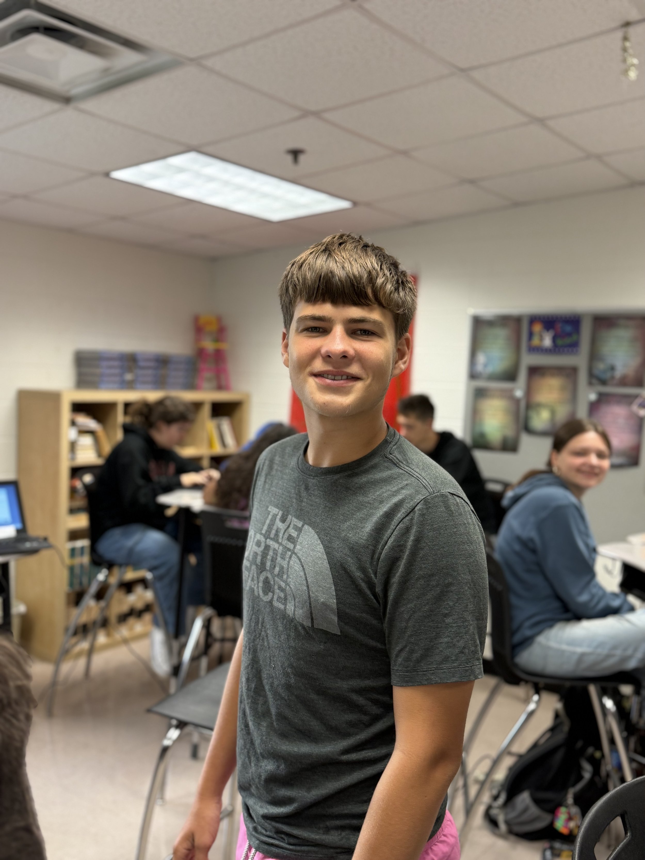 A teenage boy standing in a classroom with other students seated at desks and working on laptops in the background.