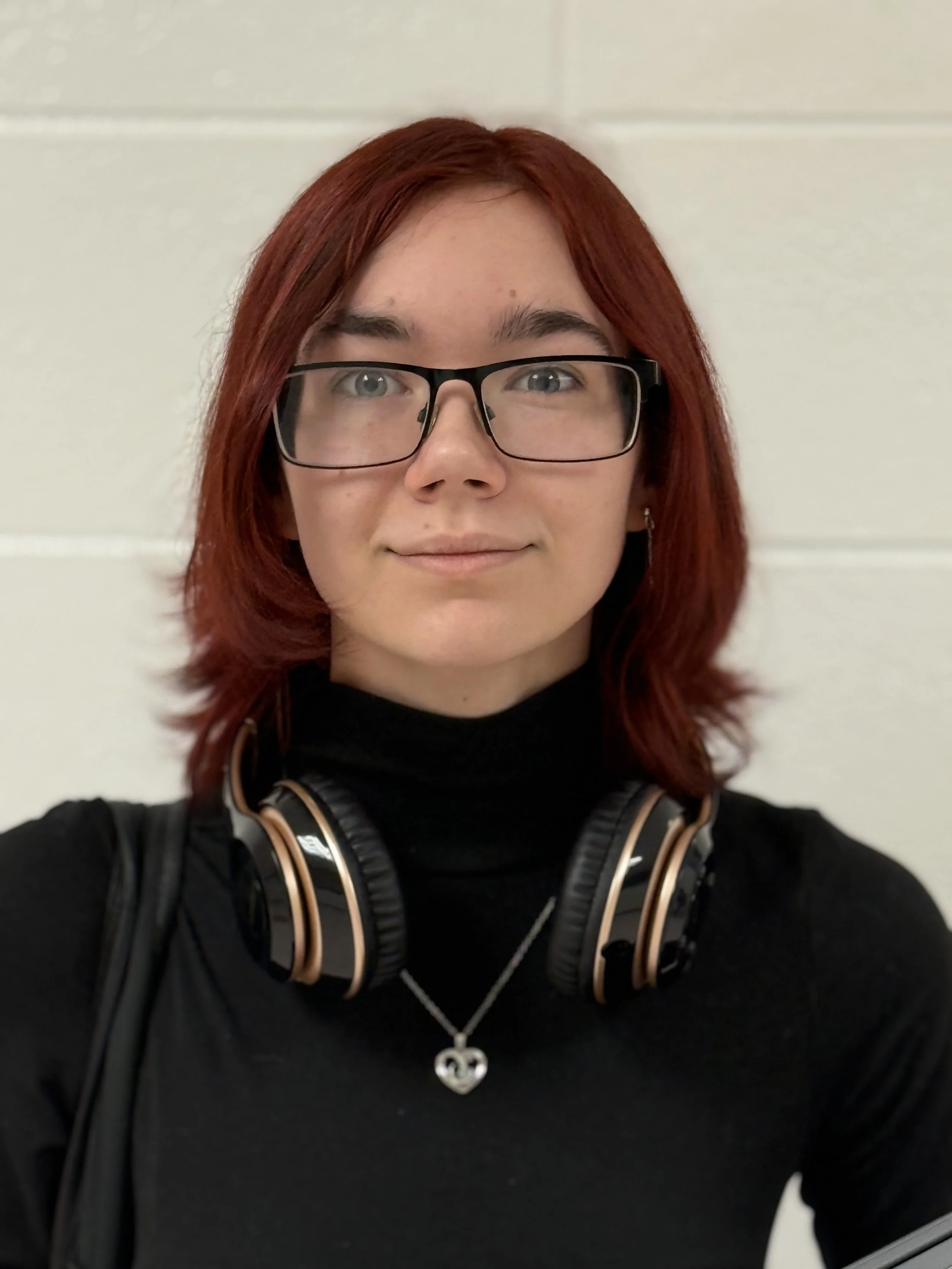 A young woman with shoulder-length red hair and glasses, wearing a black turtleneck and a necklace with a heart pendant, standing against a light-colored wall.