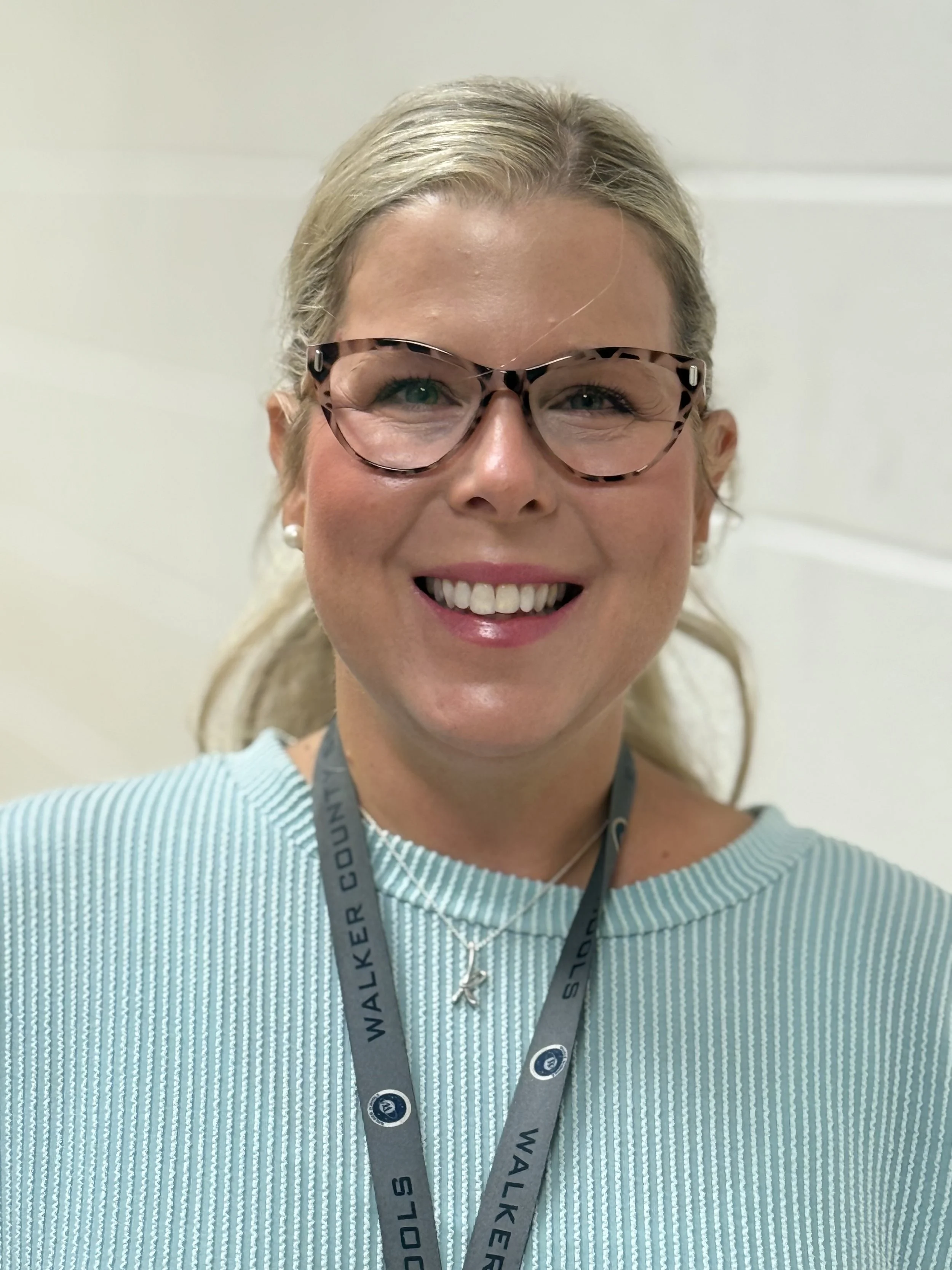 A woman with blonde hair, glasses, and a bright smile wearing a light blue striped top and a lanyard, standing against a plain background.