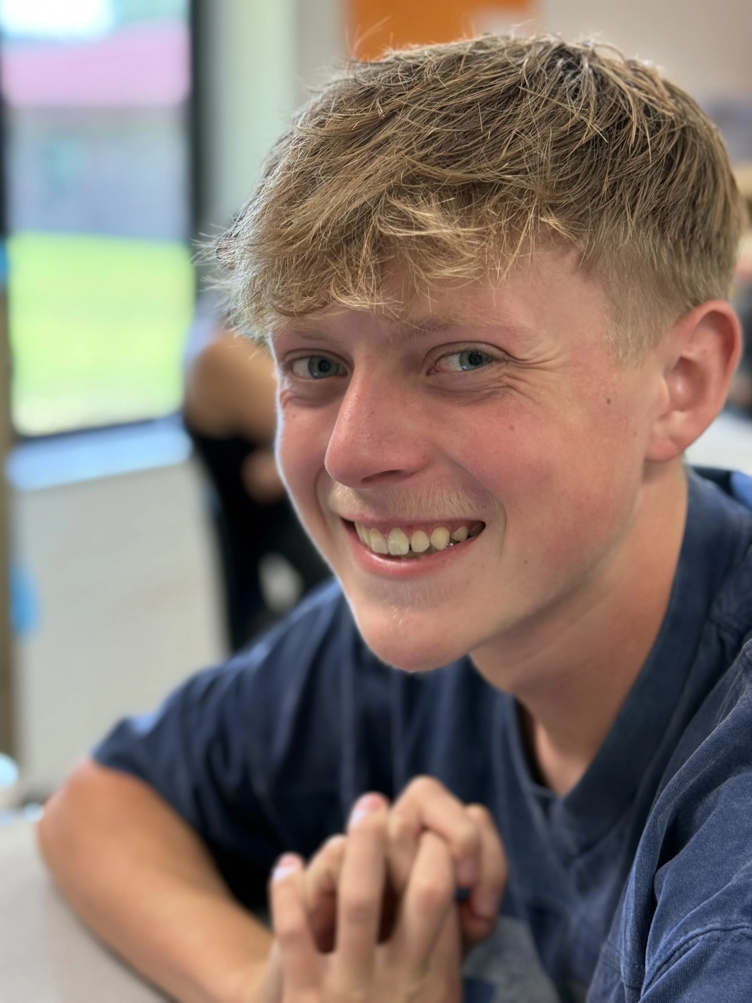 Young man with blond hair and blue eyes smiling and looking at the camera, sitting indoors with a window in the background.