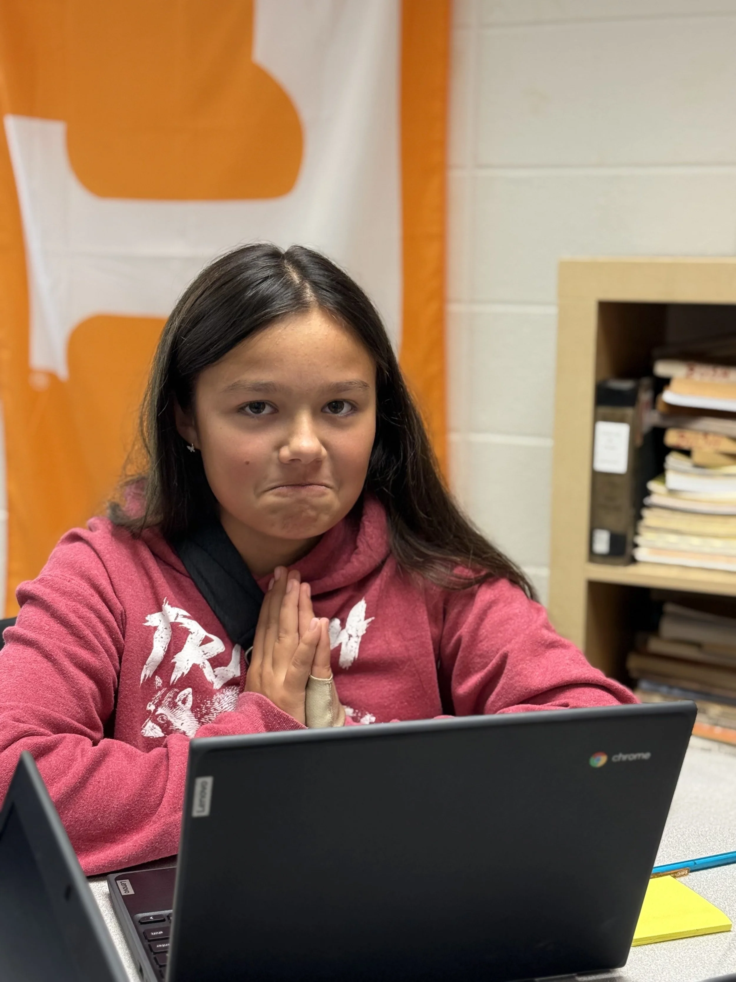 A girl with long dark hair sitting at a desk with a Chromebook. She has her hands pressed together in front of her chest and an expression that suggests she is praying or hoping. She is wearing a maroon hoodie.