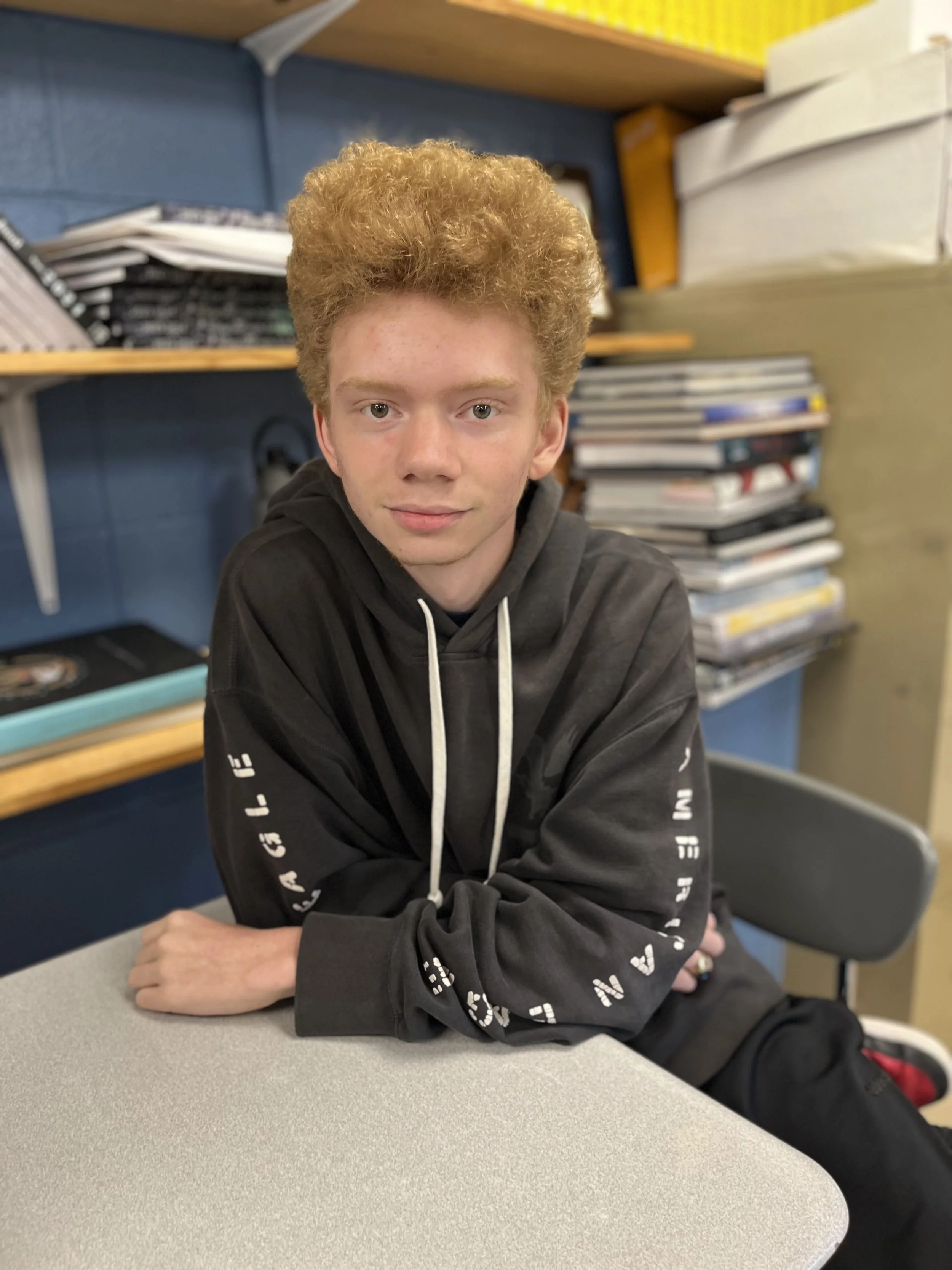 A young person with curly red hair and light skin, smiling in a classroom with a colorful poster or book in the background.