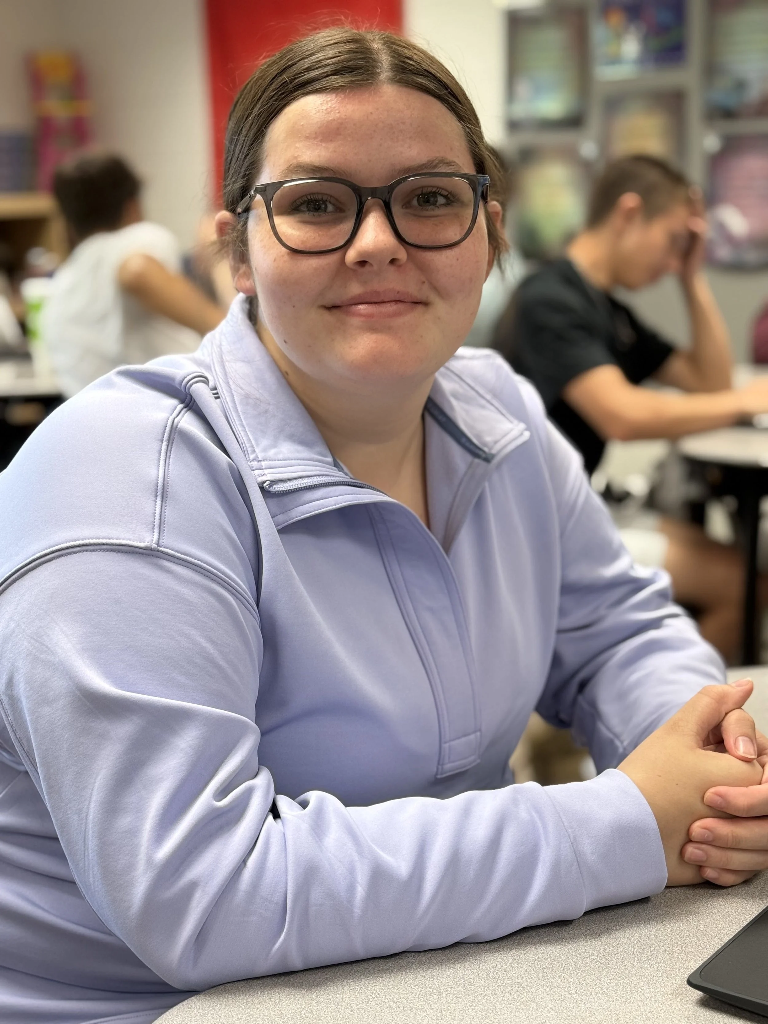 A young woman with glasses and a light purple zip-up jacket sitting at a desk in a classroom, smiling at the camera. In the background, other students are working and talking.