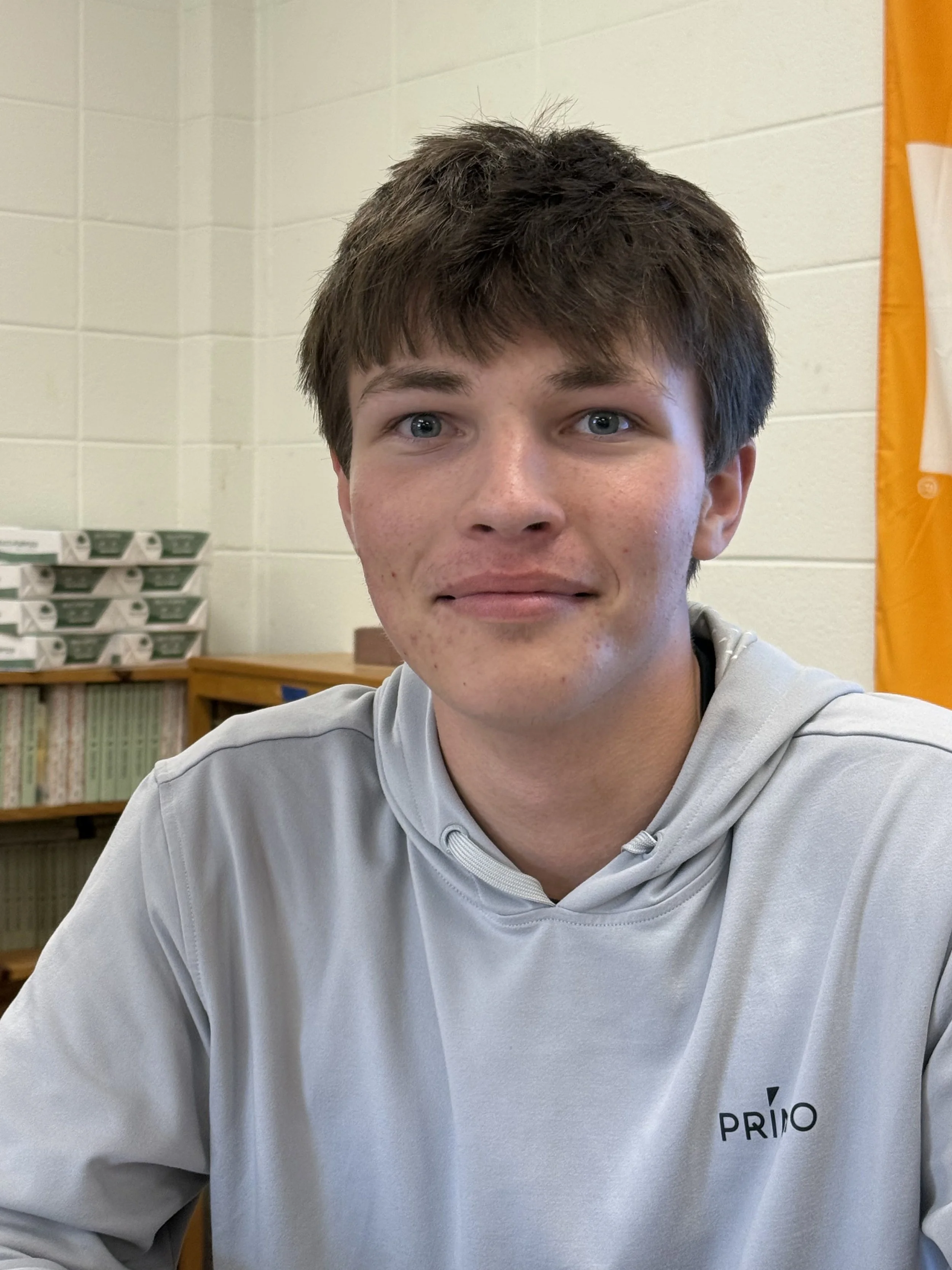 A teenage boy sitting at a desk in a room with beige cabinets and a window. He is wearing a white t-shirt and looking at the camera with a slight smile. There is a water bottle on the desk in front of him.