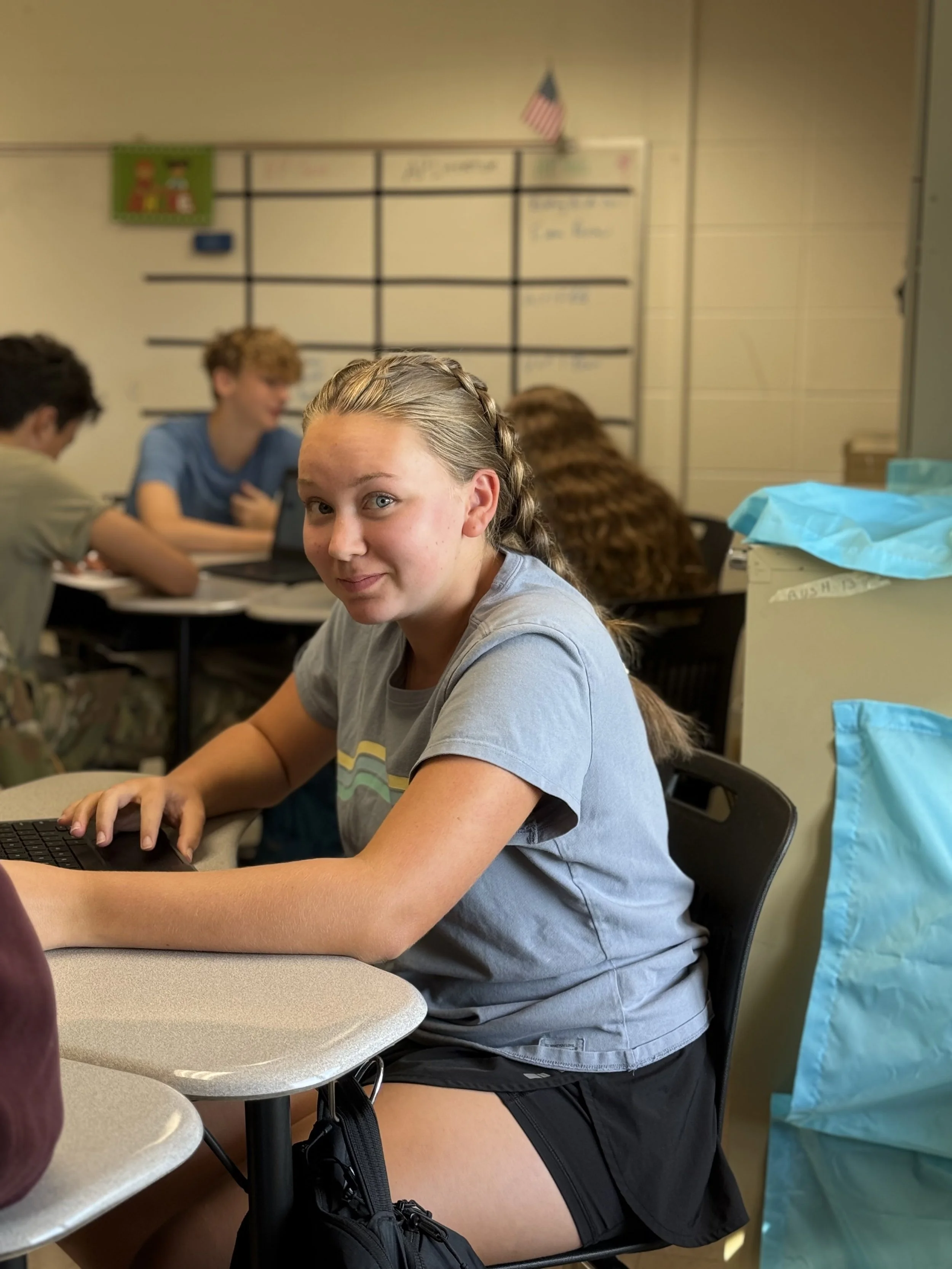 A teenage girl with braided hair sitting at a classroom desk, using a laptop, with other students in the background.