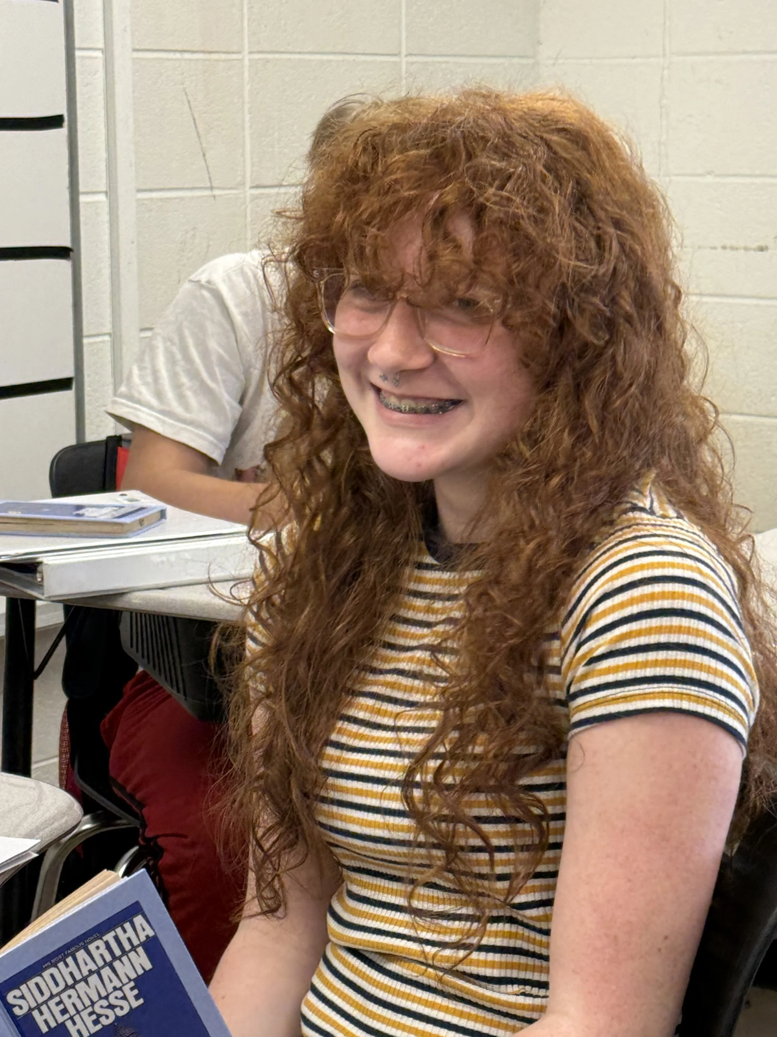A young woman with red hair in braids, light skin, and a septum piercing, sitting at a desk with lined paper and pink highlighter, resting her chin on her hand.