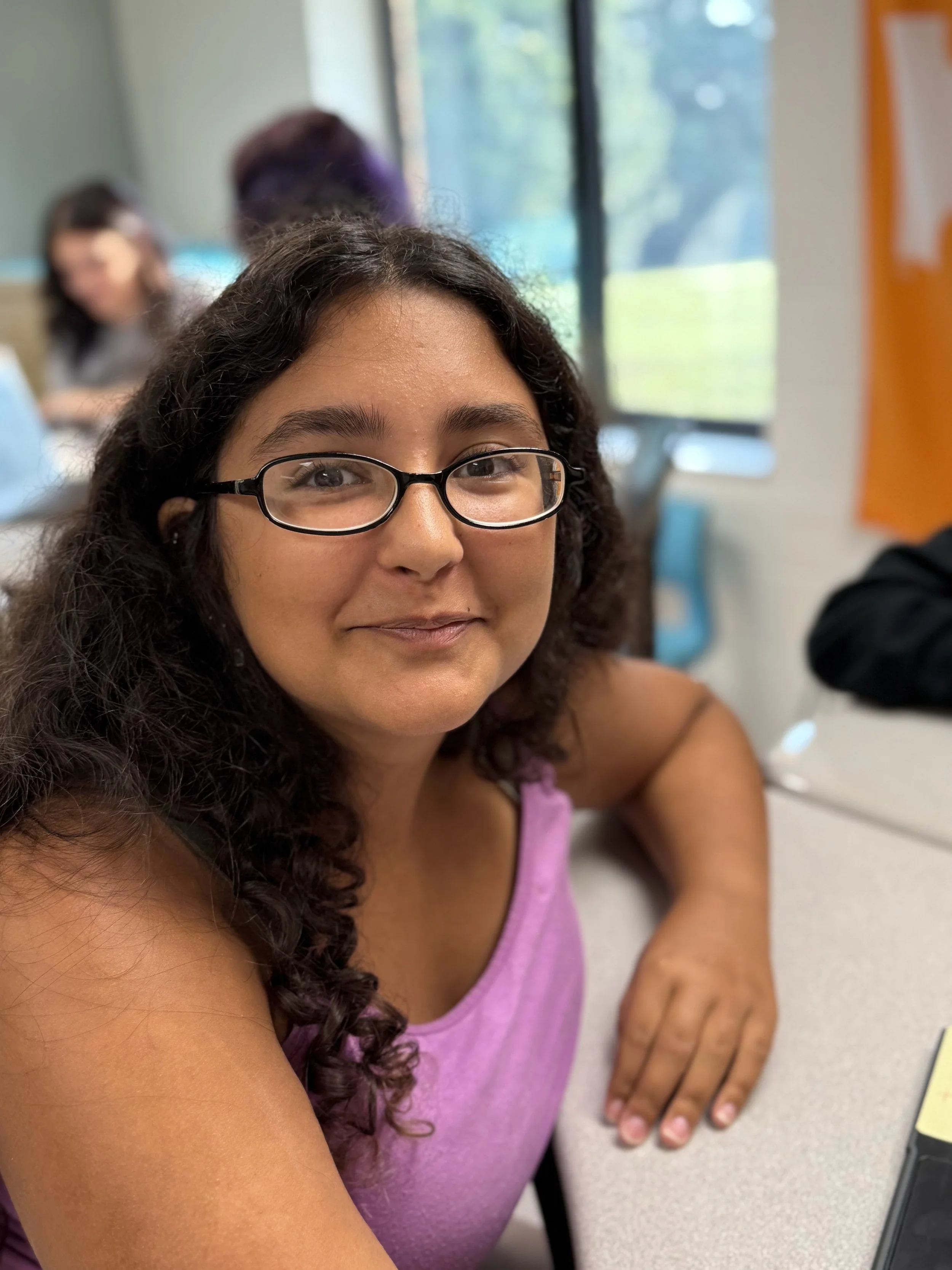 A young woman with glasses and curly hair, smiling at the camera, sitting at a desk in a classroom with other students and large windows in the background.