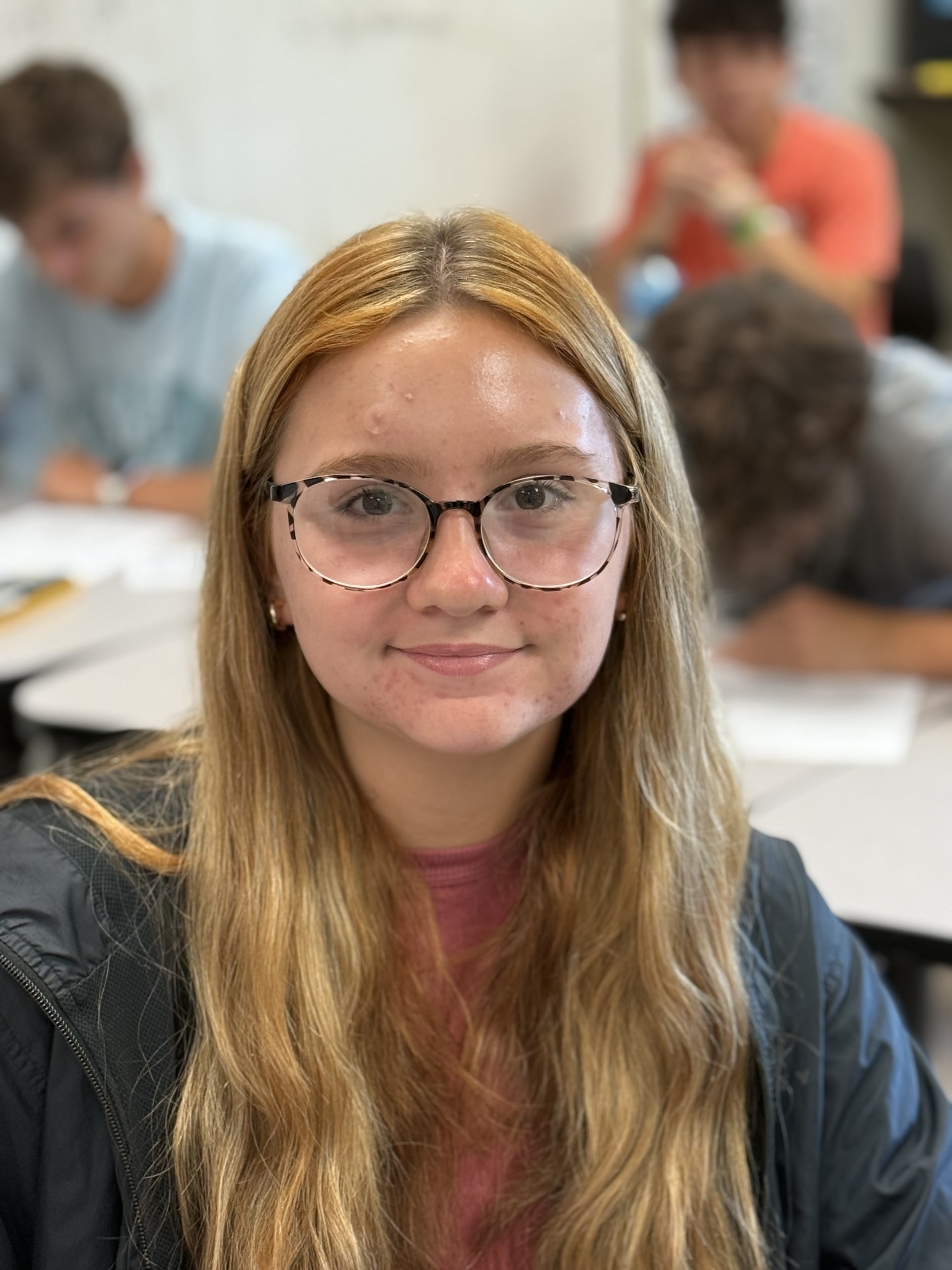 A young woman with long blonde hair, glasses, and a pink shirt, sitting at a table in a classroom with other students in the background.