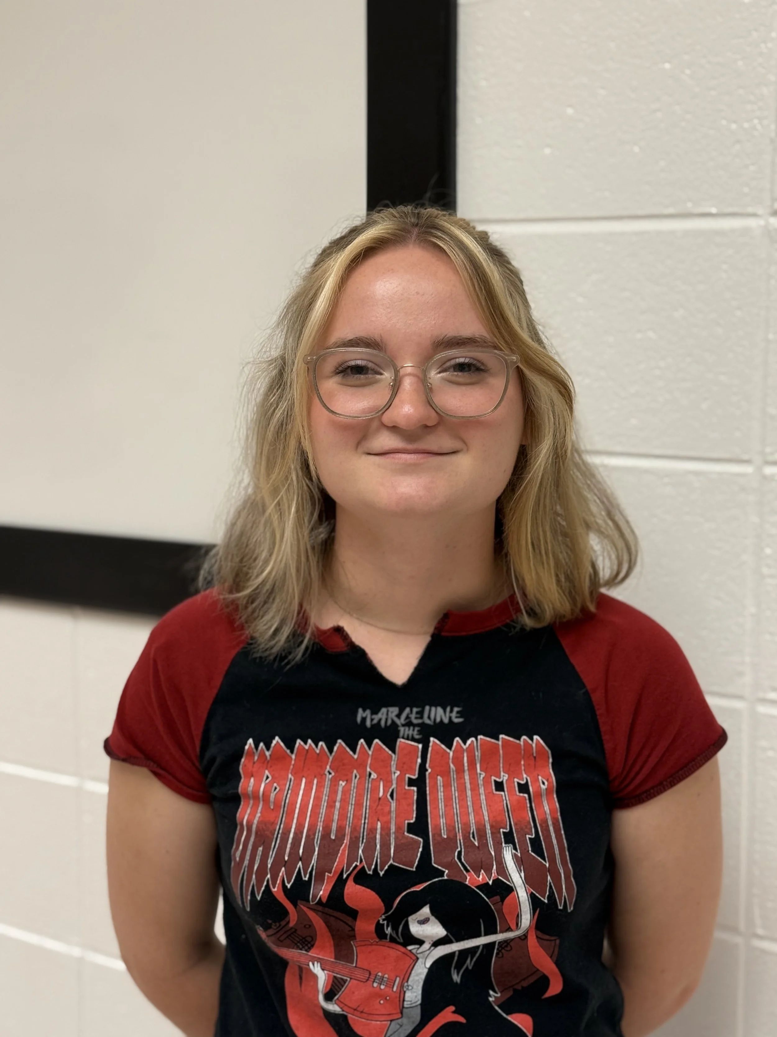 A young woman with shoulder-length blonde hair, glasses, and a maroon and black graphic t-shirt standing indoors against a white wall.