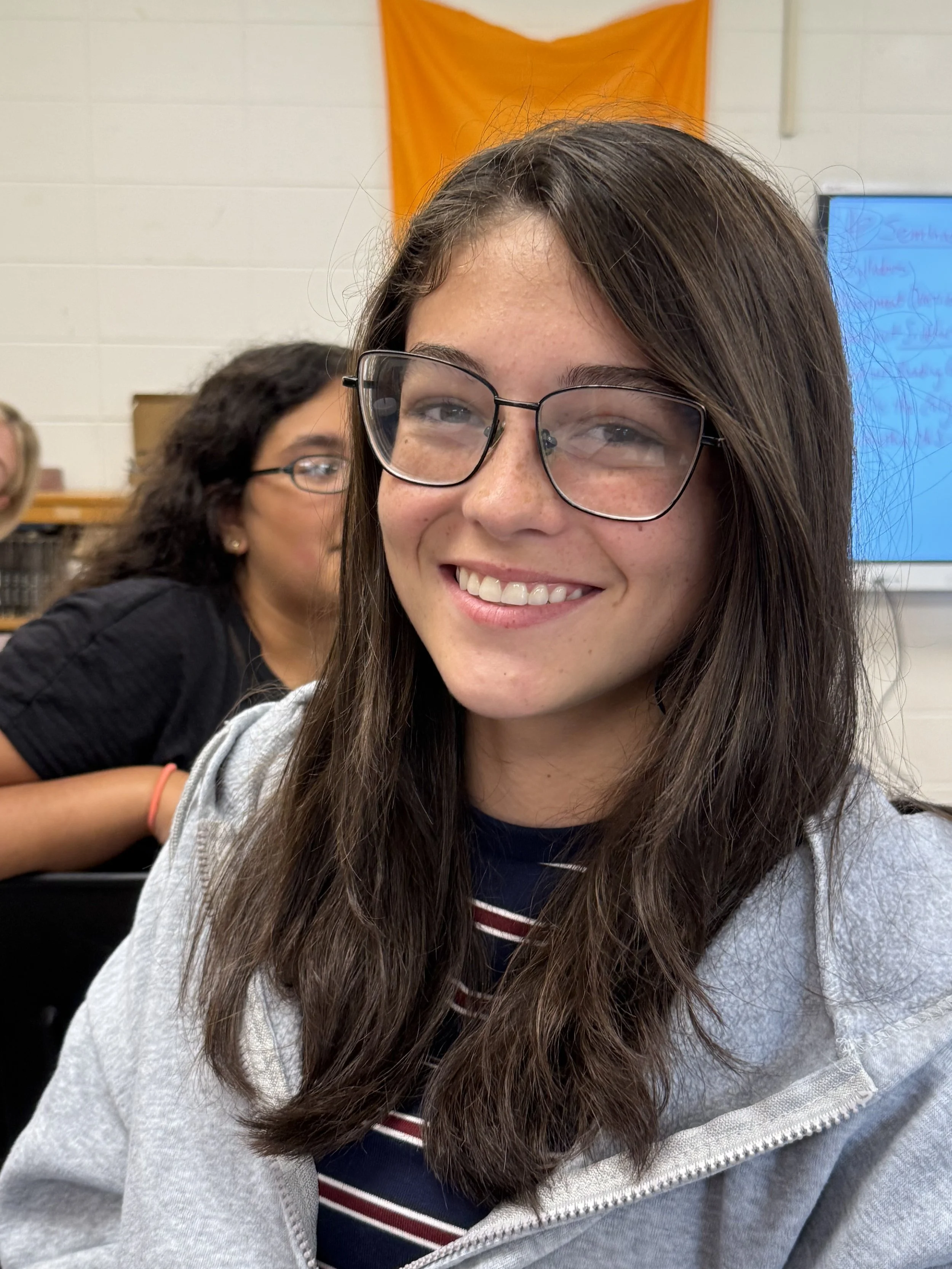 Close-up of a young woman with long, wavy dark hair wearing glasses, smiling at the camera, in a classroom setting.