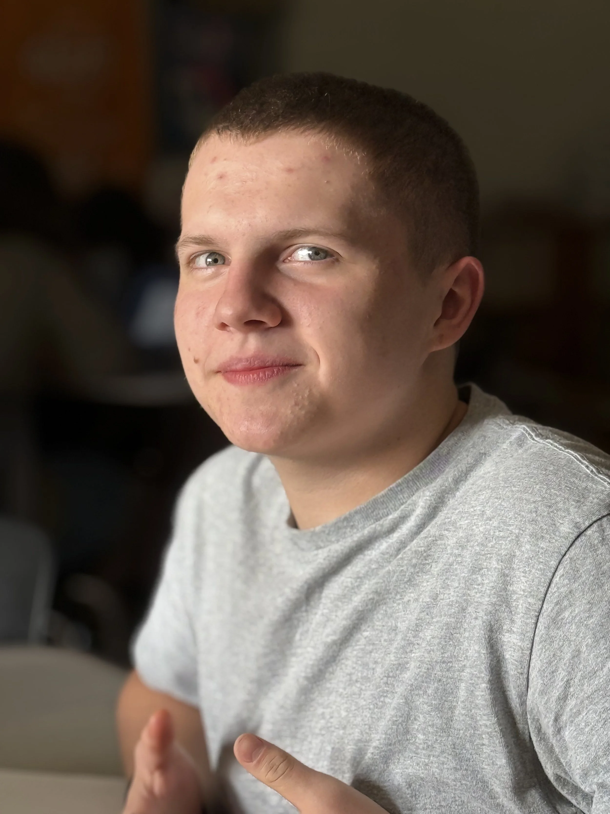 A young man with short hair, light skin, and blue eyes looking at the camera with a slight smile, wearing a gray t-shirt with a blurred indoor background.