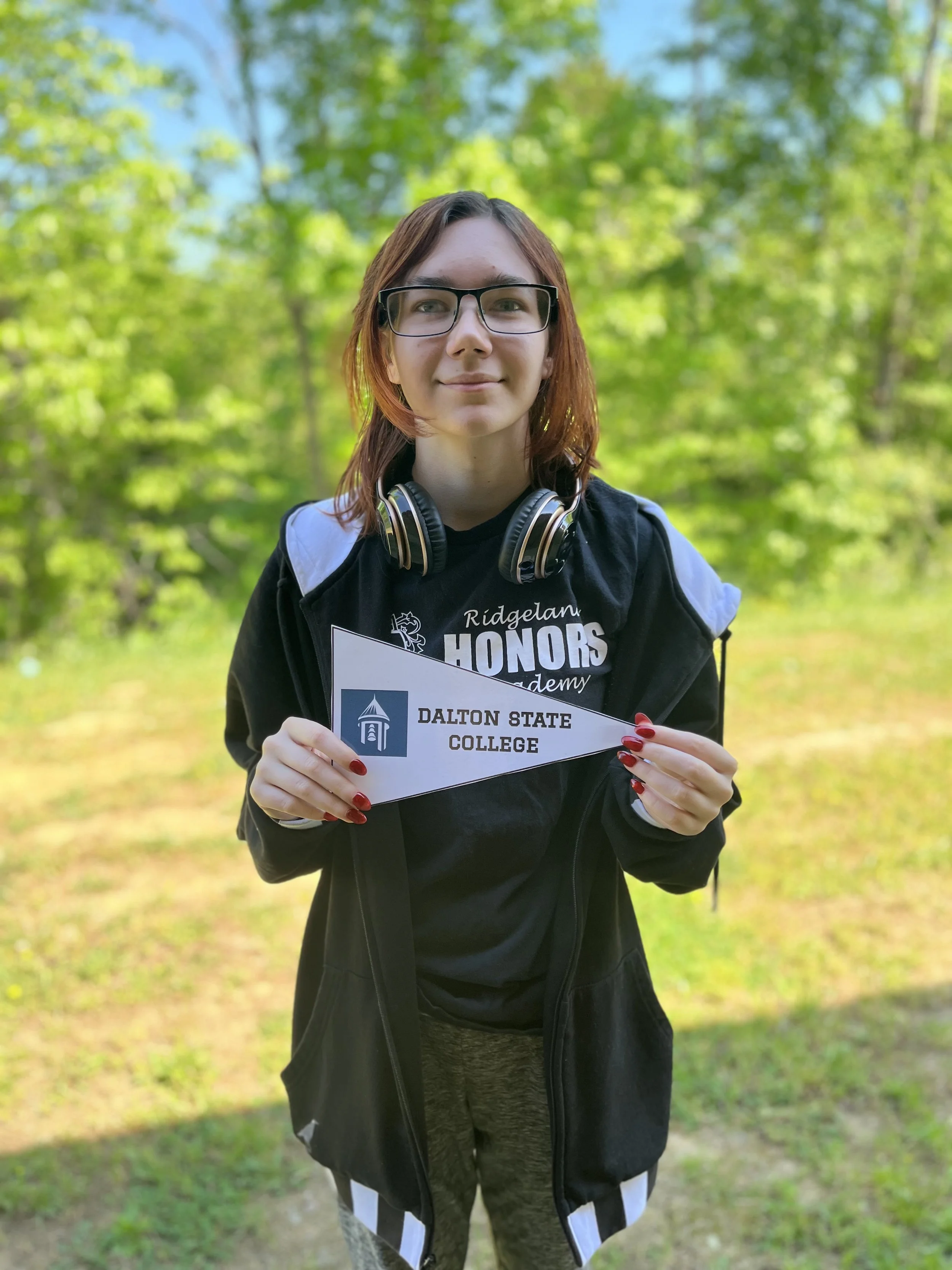 A young woman with shoulder-length red hair and glasses, wearing a black turtleneck and a necklace with a heart pendant, standing against a light-colored wall.