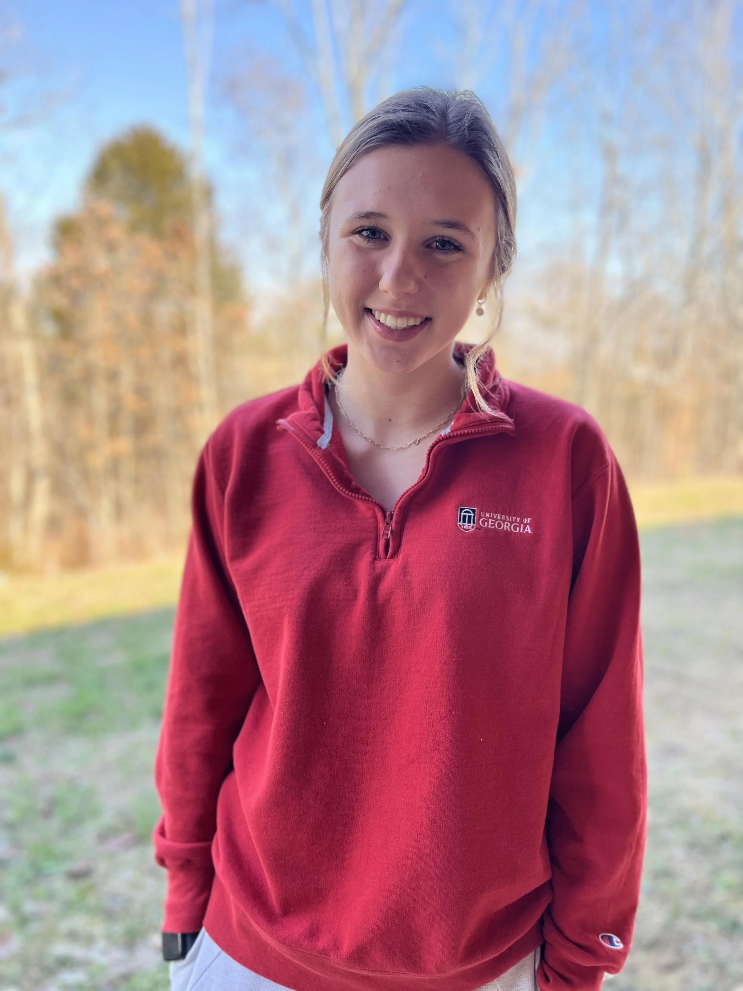 A young woman with light brown hair, smiling, wearing a pink shirt and a dark jacket, sitting in a room with orange chairs in the background.