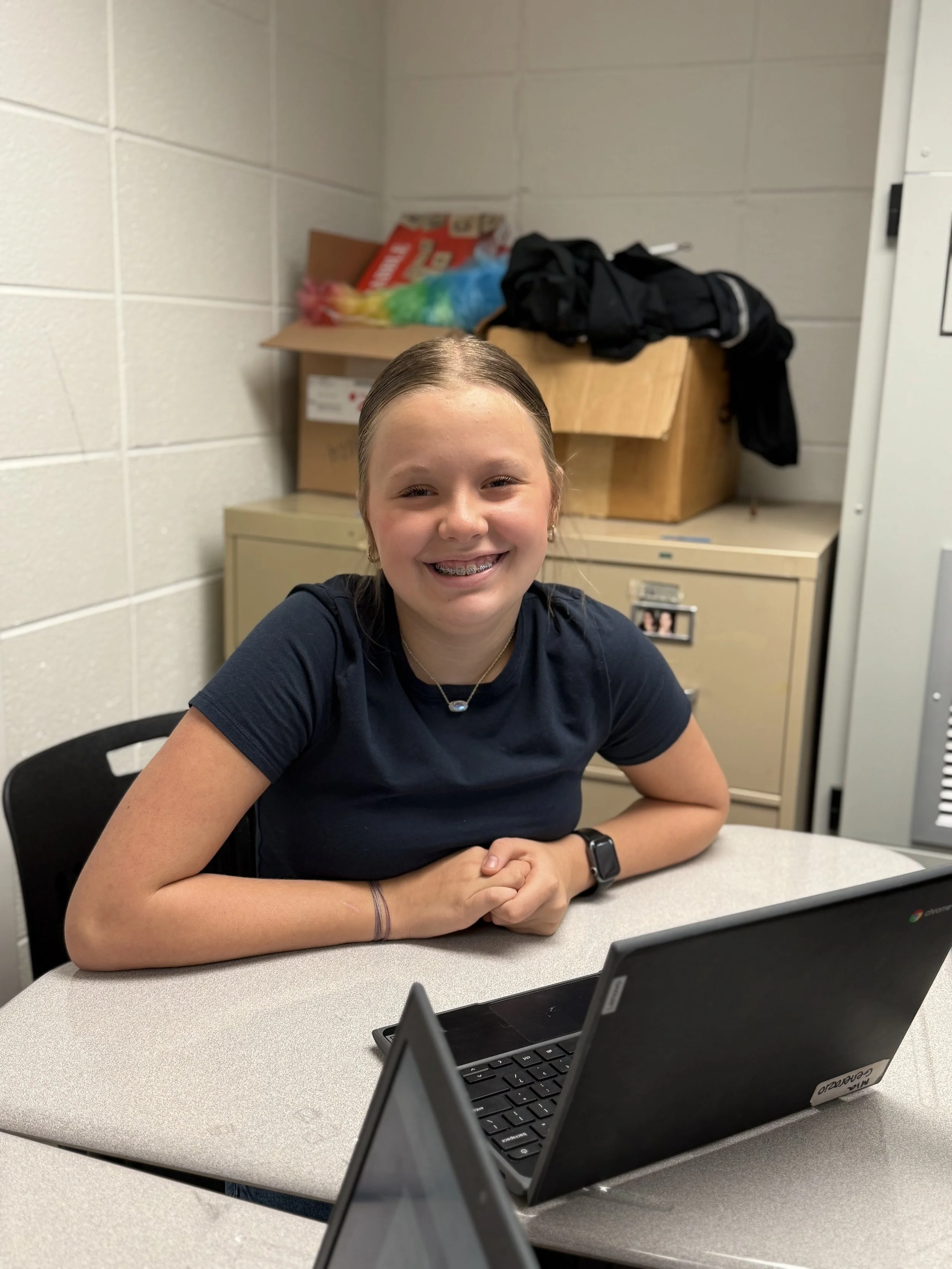 A smiling girl with braces sitting at a table in a classroom or office, with a Chromebook in front of her and a beige filing cabinet behind her.