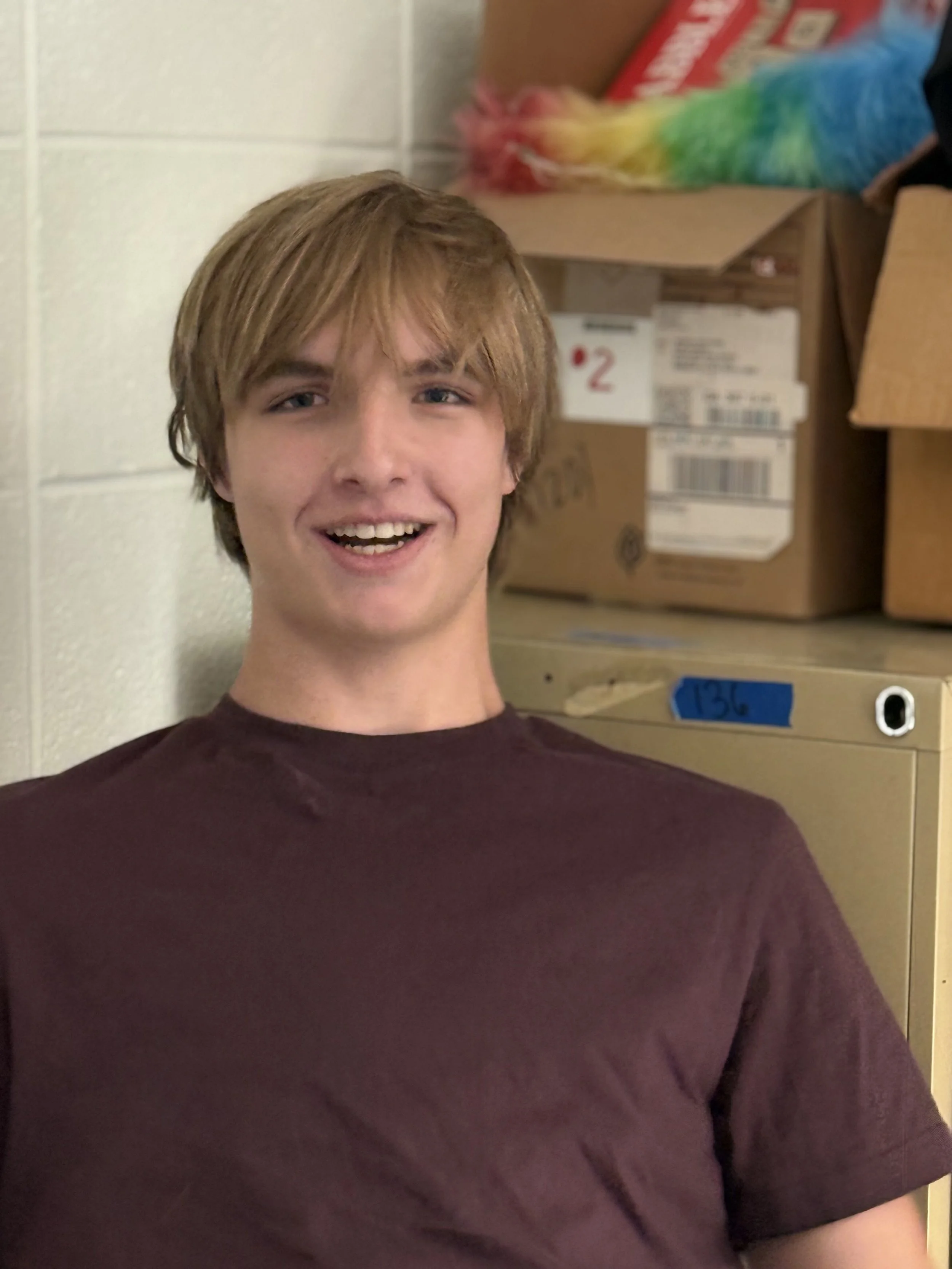 A young man with light brown hair, smiling, wearing a maroon T-shirt, standing in a room with boxes and a rainbow-colored plush item in the background.