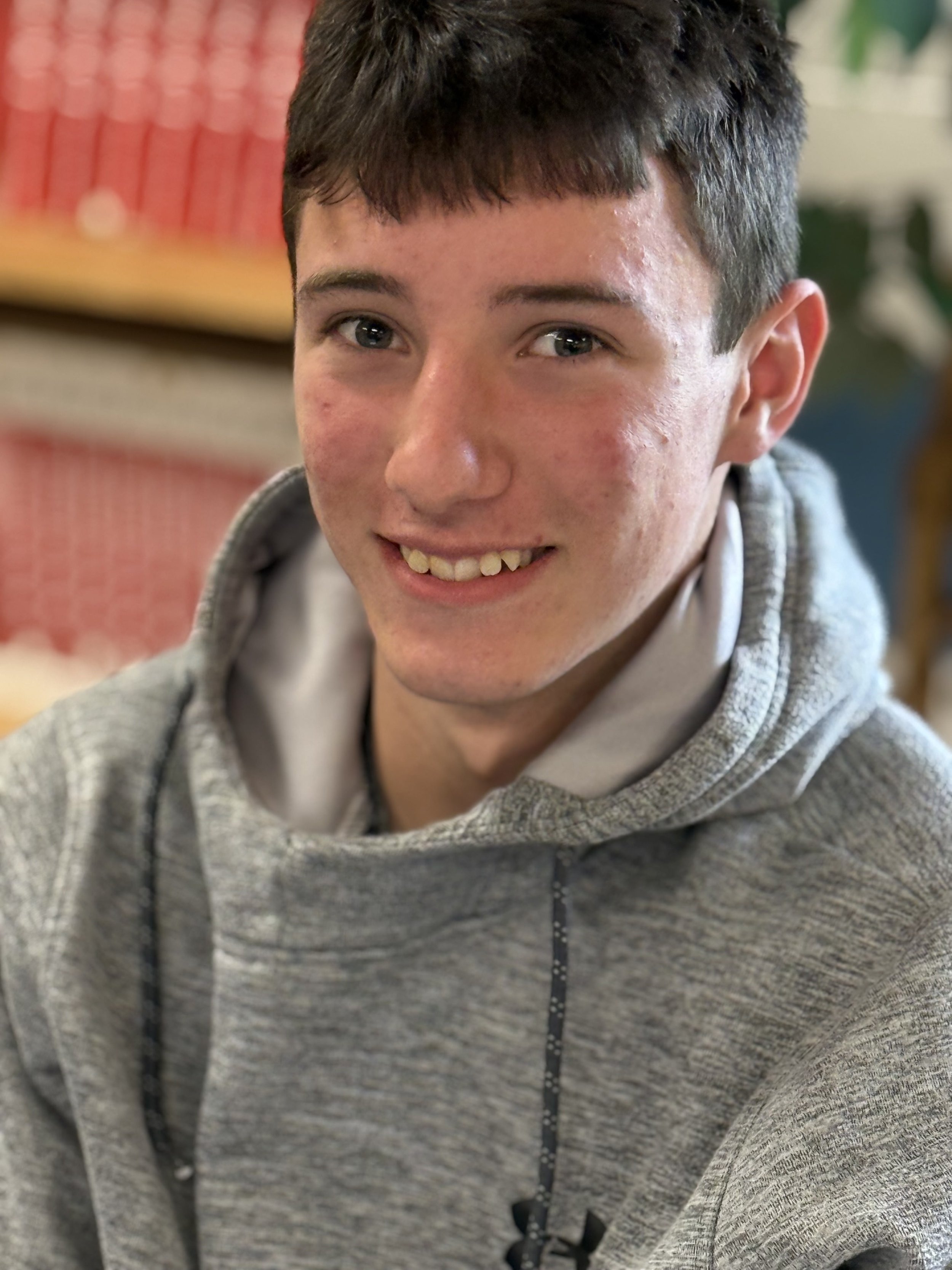 A teenage boy with short dark hair smiling, wearing a grey hoodie with a drawstring.
