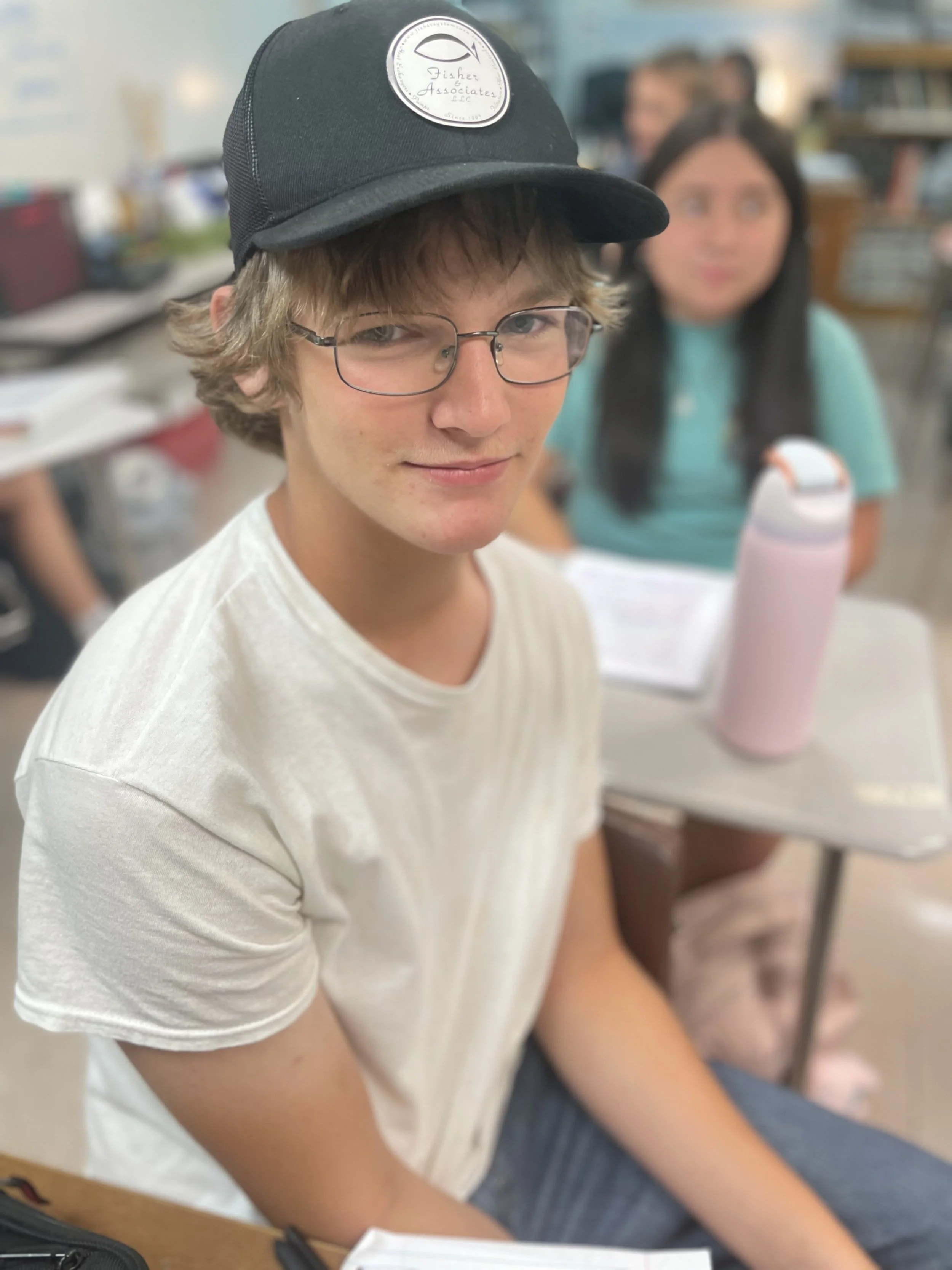 A teenage boy with curly light brown hair and glasses, wearing a black baseball cap and a white t-shirt, sitting at a table in a busy indoor setting.