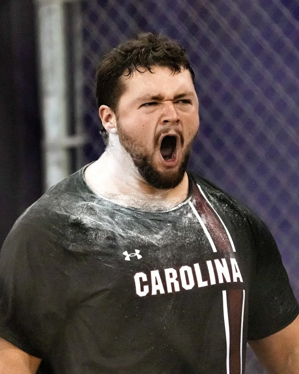 A man with short curly hair and a beard, wearing a black Carolina sports shirt, is mid-shout or roar with his mouth wide open, and white powder dusted on his neck and upper chest.