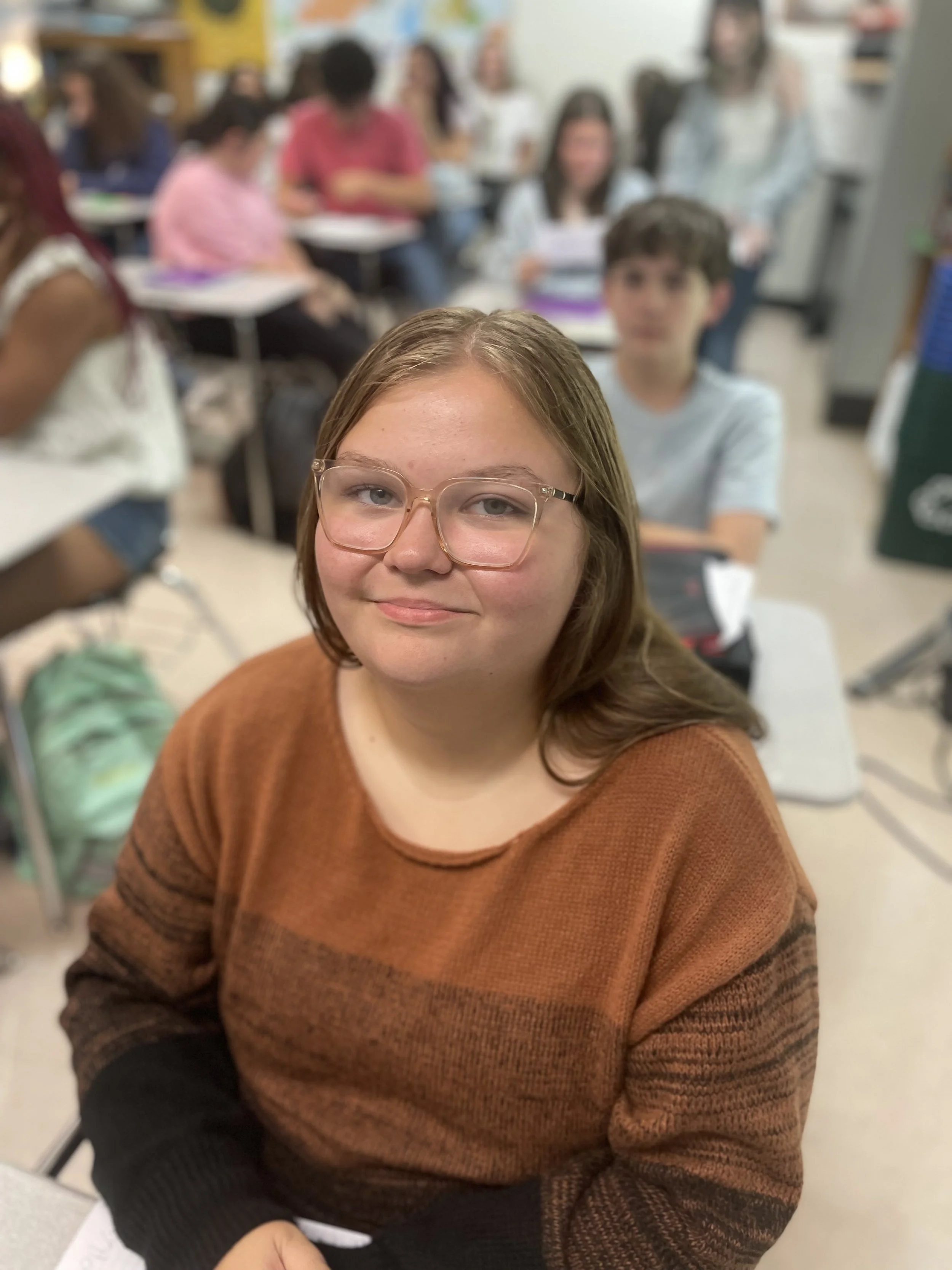 A girl with glasses and brown hair sitting at a desk in a classroom, smiling at the camera with students and teacher in the background.