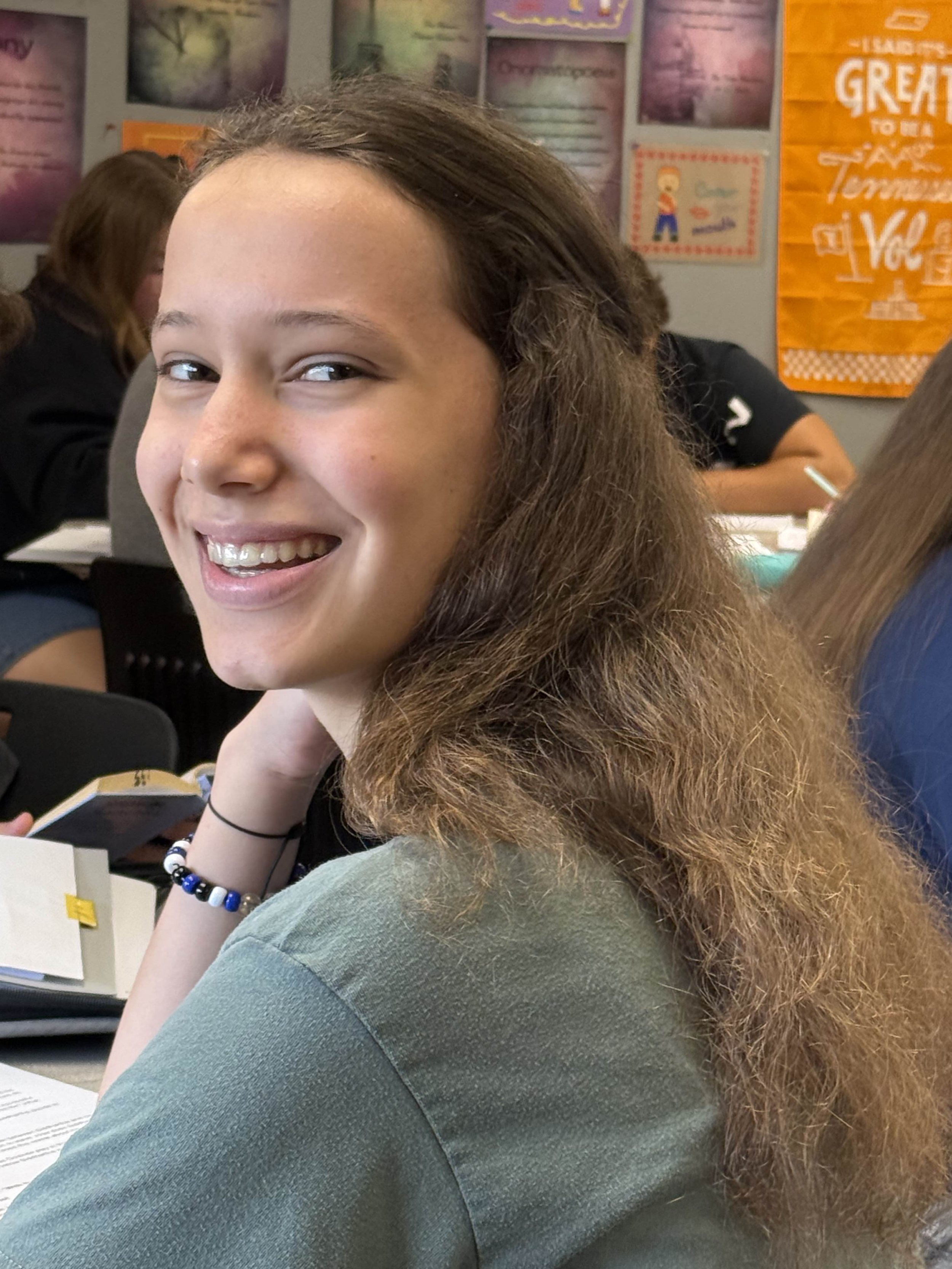 A smiling girl with braces sitting at a desk in a classroom, wearing a blue hoodie with her arms crossed on the table. There are notebooks and pens on the desk.