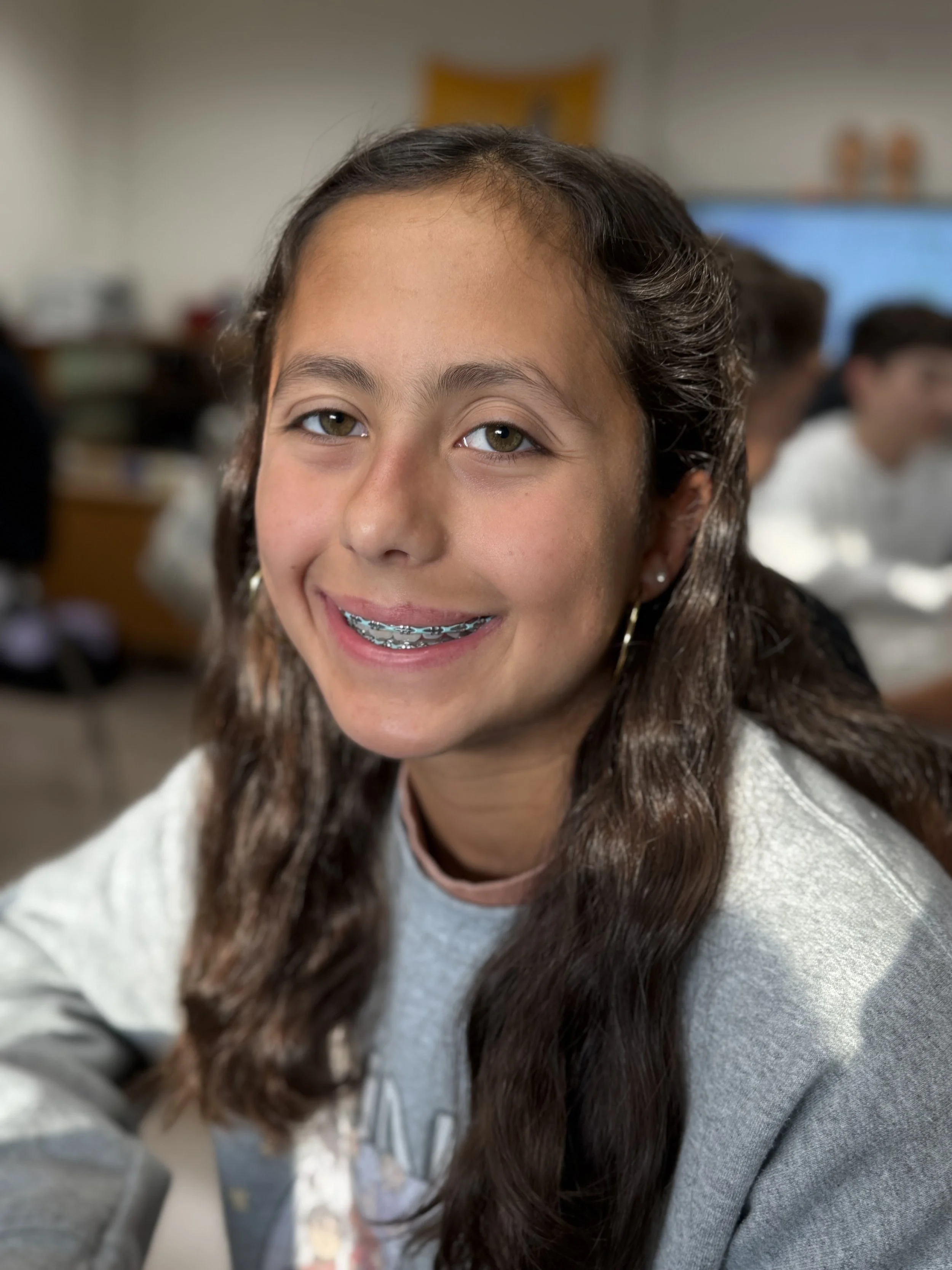 Smiling girl with braces and long brown hair in a classroom.