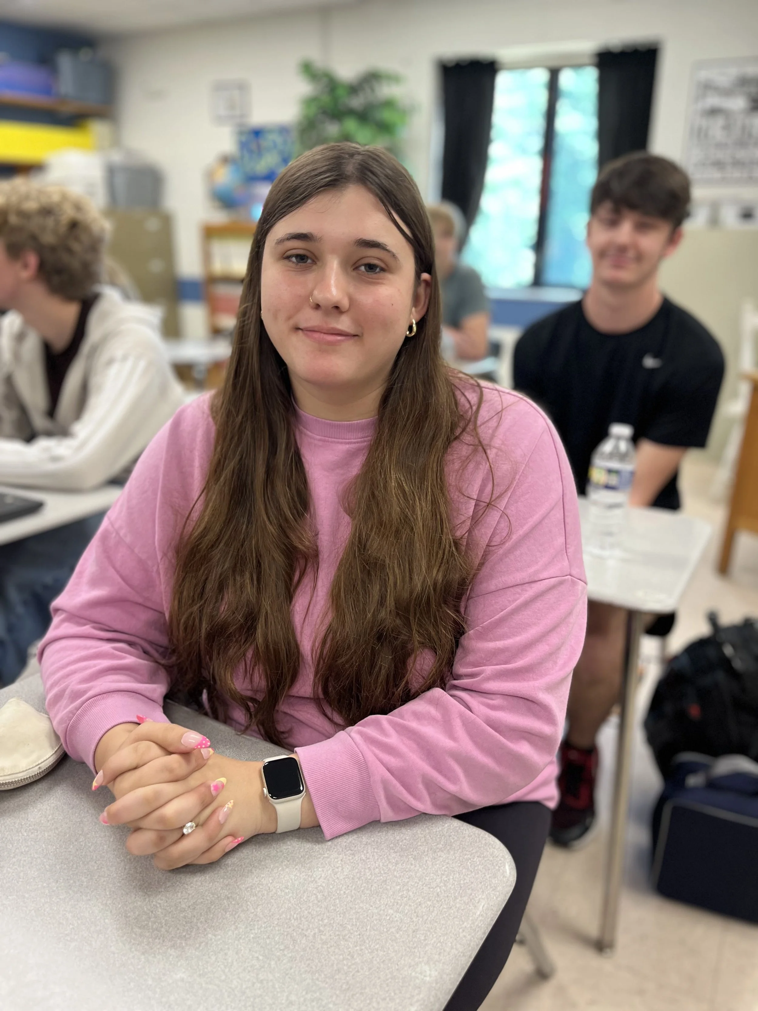 A young woman with long brown hair and a nose piercing, wearing a pink shirt, sitting at a desk in a classroom.
