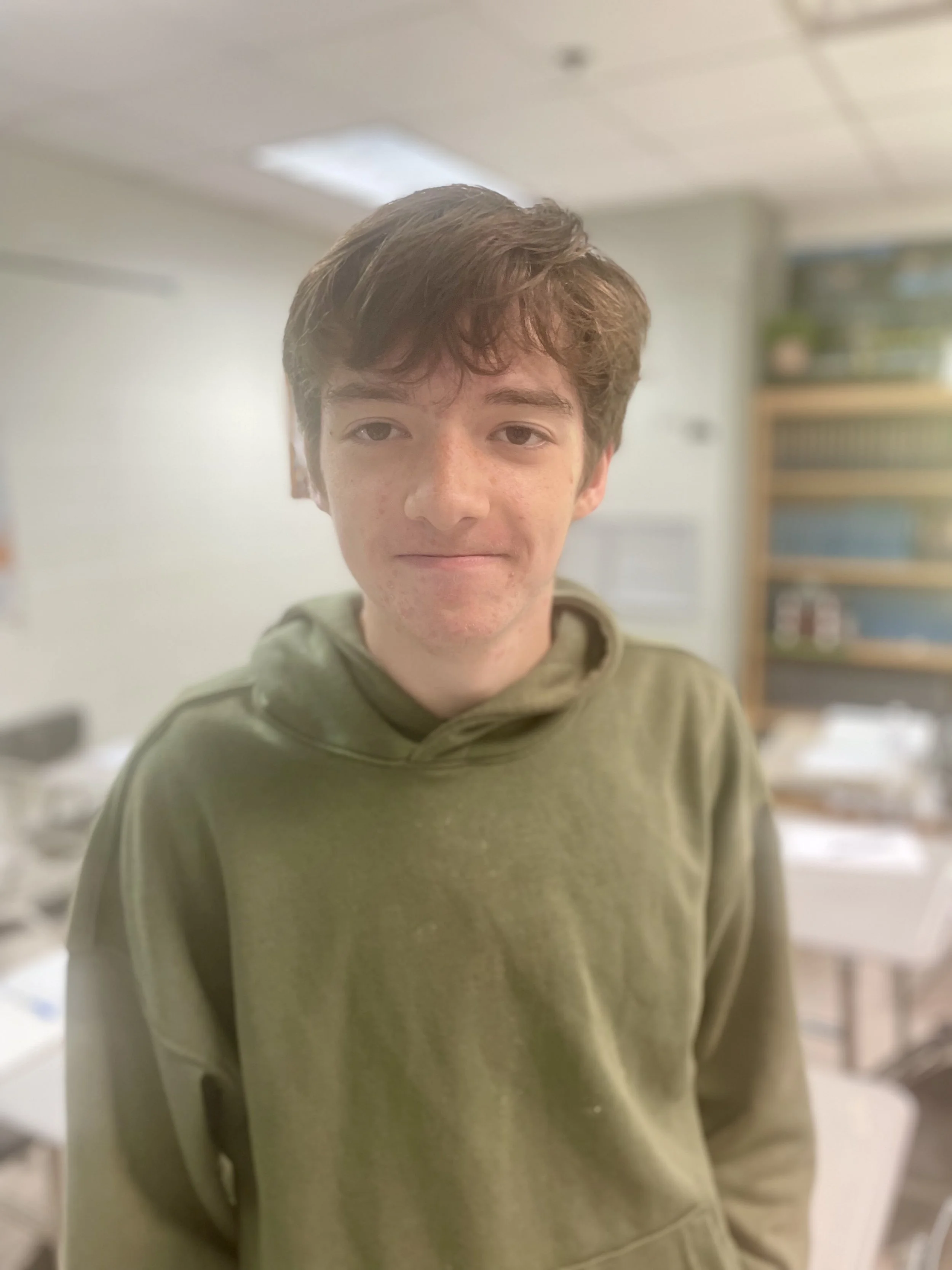 A young man with wavy brown hair, wearing a green hoodie, standing indoors with a blurred background of tables and shelves.
