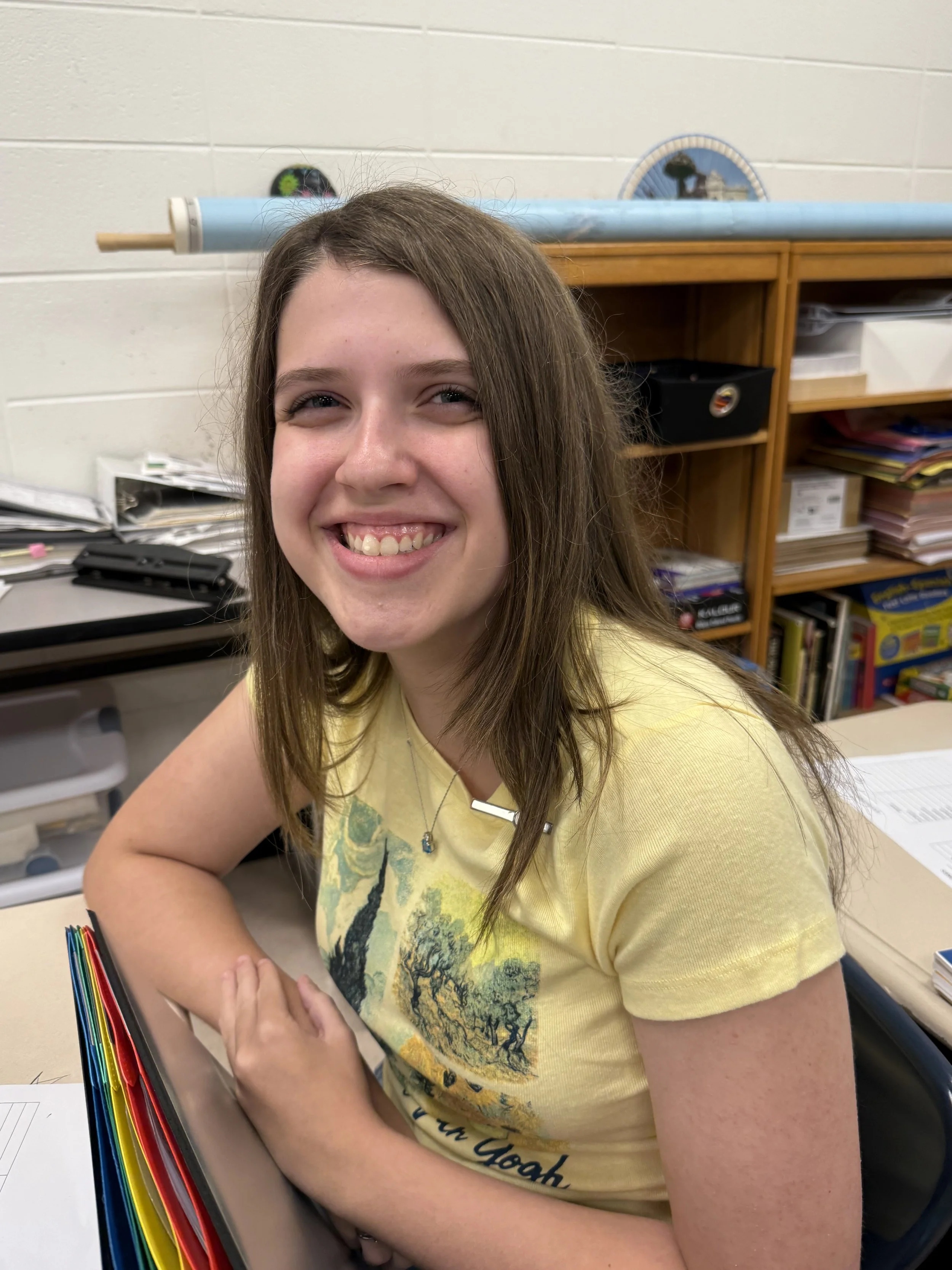 A smiling girl with long light brown hair sitting at a desk in a classroom, wearing a light-colored t-shirt with strawberries and smiling at the camera.