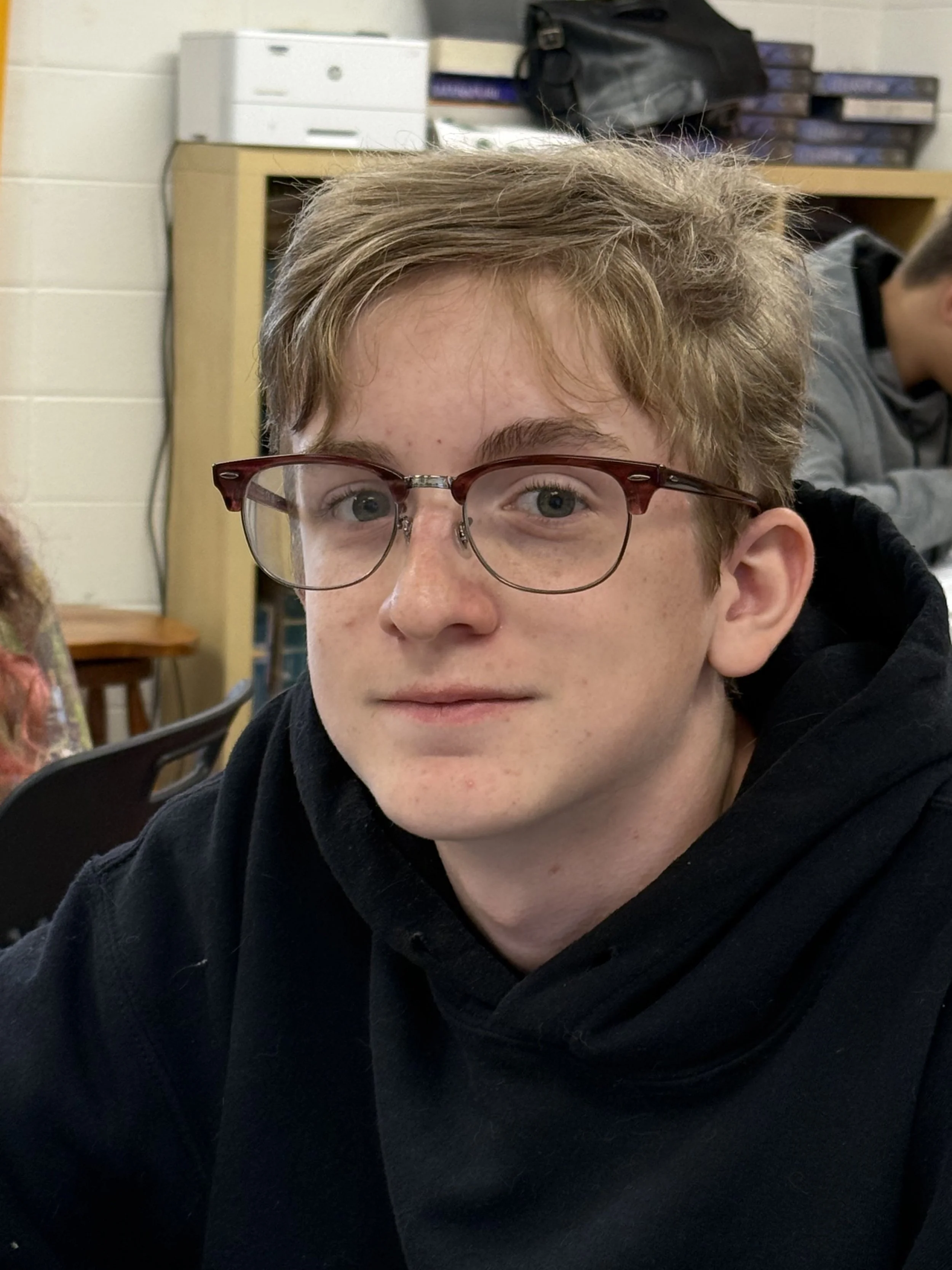 A young boy with red glasses and light brown hair wearing a blue and red jacket sits at a desk in a classroom, with other students in the background.