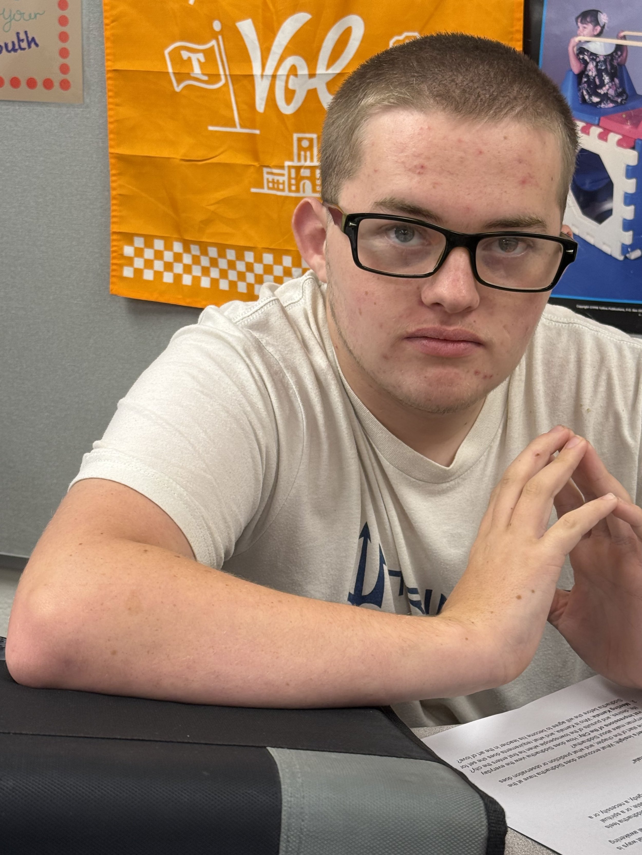 A young man with glasses sitting at a table in front of a laptop, wearing a Vanderbilt hoodie, with a red and gold banner behind him that has the Arsenal logo.