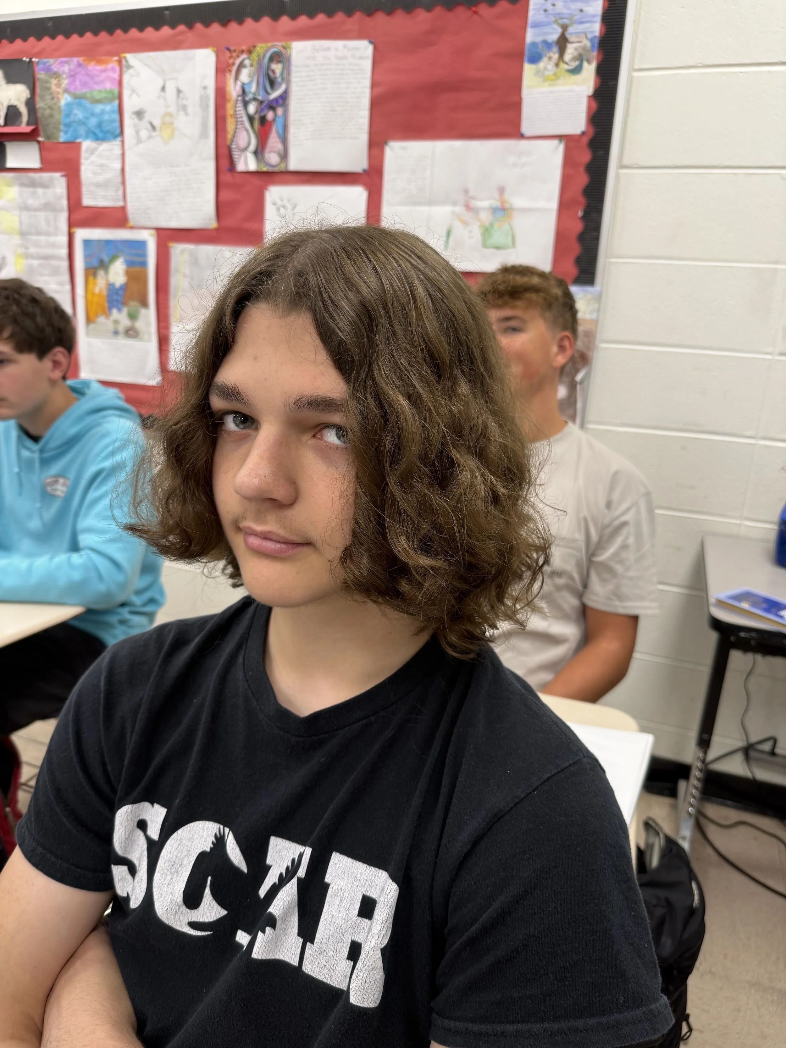 A teenage girl with curly brown hair sitting at a desk, resting her chin on her hands with a red pen held between her fingers, and looking at the camera. She is wearing a grey T-shirt, a wristwatch, and wristbands. A boy with similar hair and a blue shirt is blurred in the background.