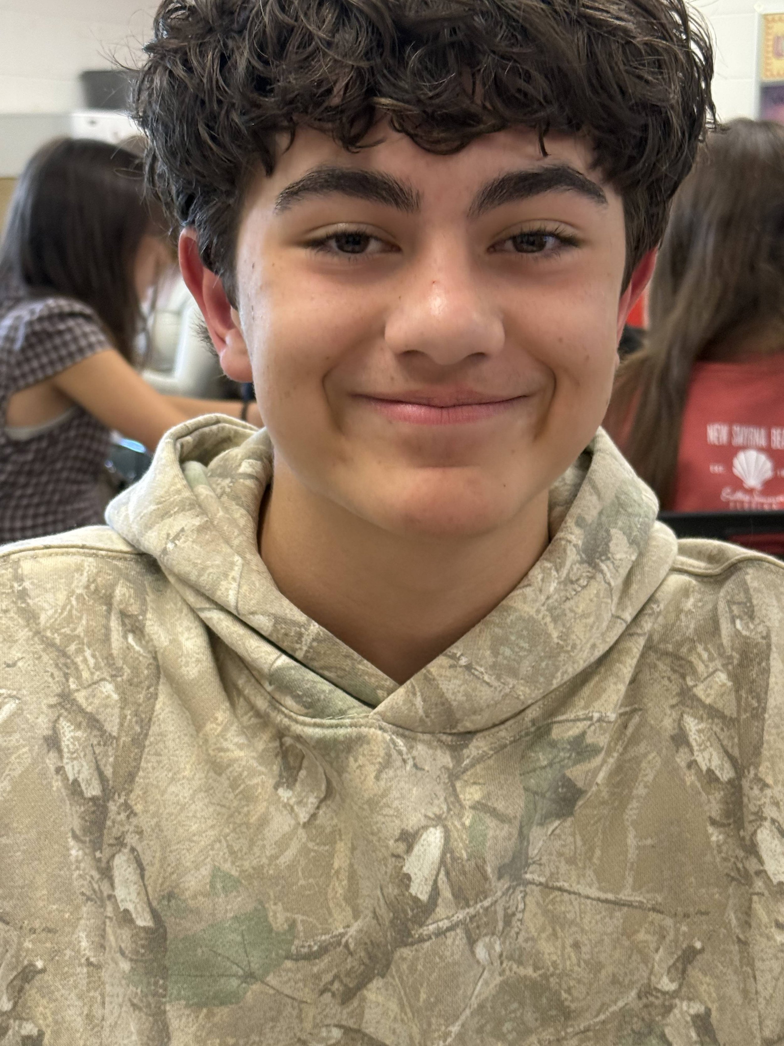 A teenage boy with brown, curly hair smiling while sitting at a table in a classroom.