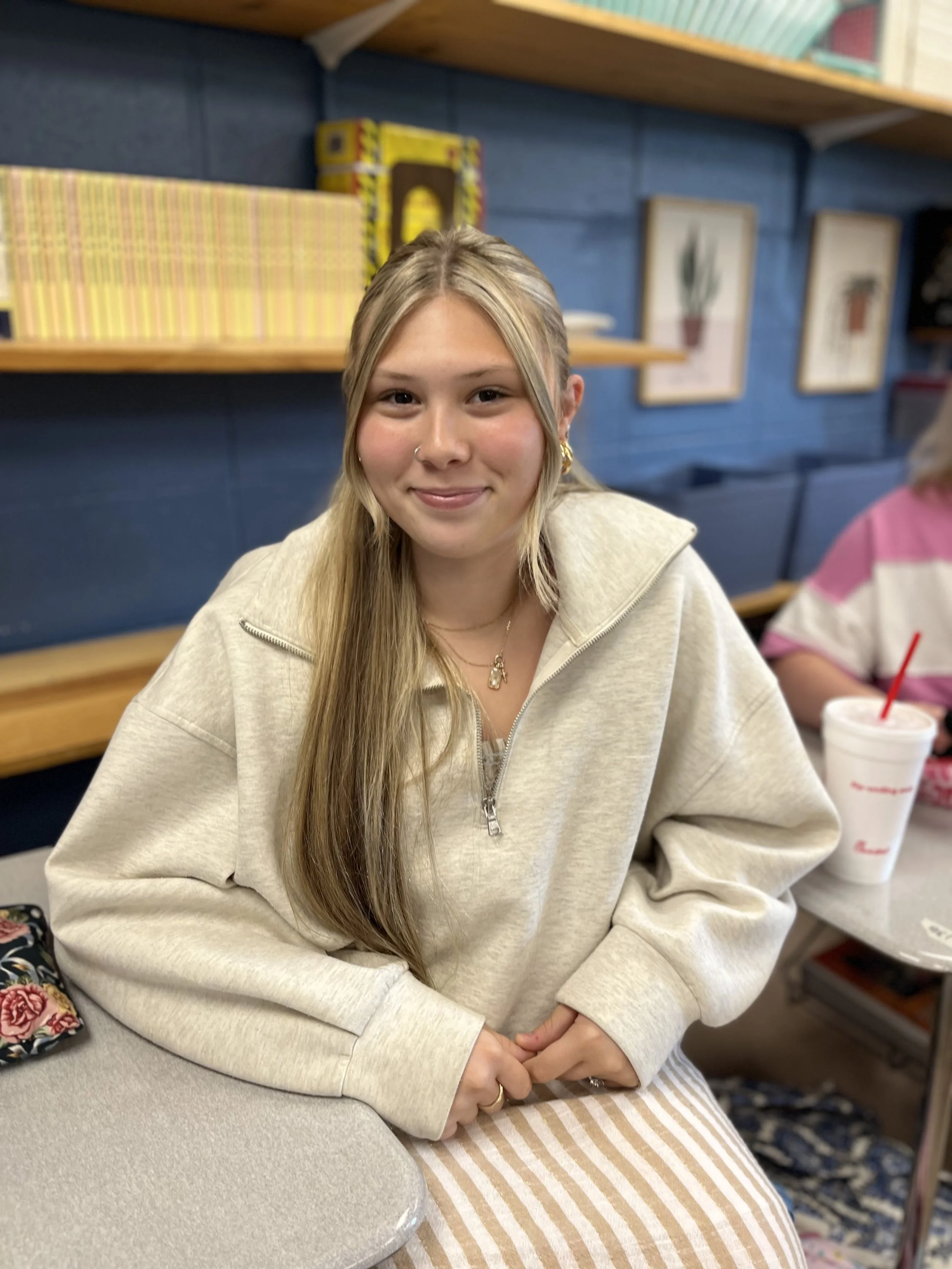 A young woman with blonde hair, wearing earrings and a necklace, smiling at the camera in an indoor setting with books or files on shelves in the background.