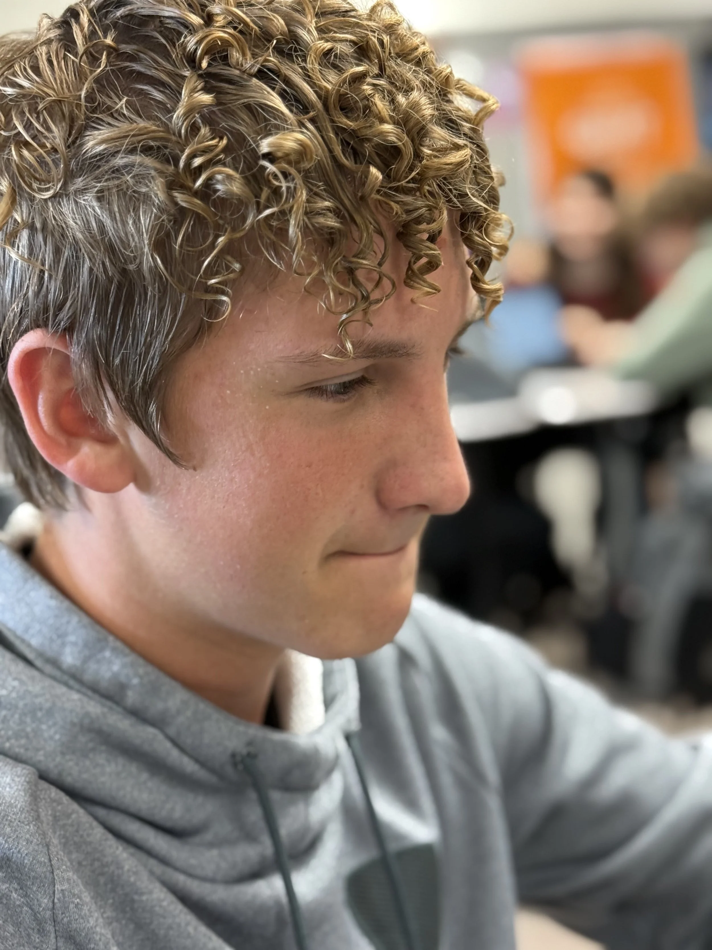 Close-up of a young man with curly blonde hair, wearing a gray hoodie, sitting indoors with a blurred background of people in a classroom or similar setting.