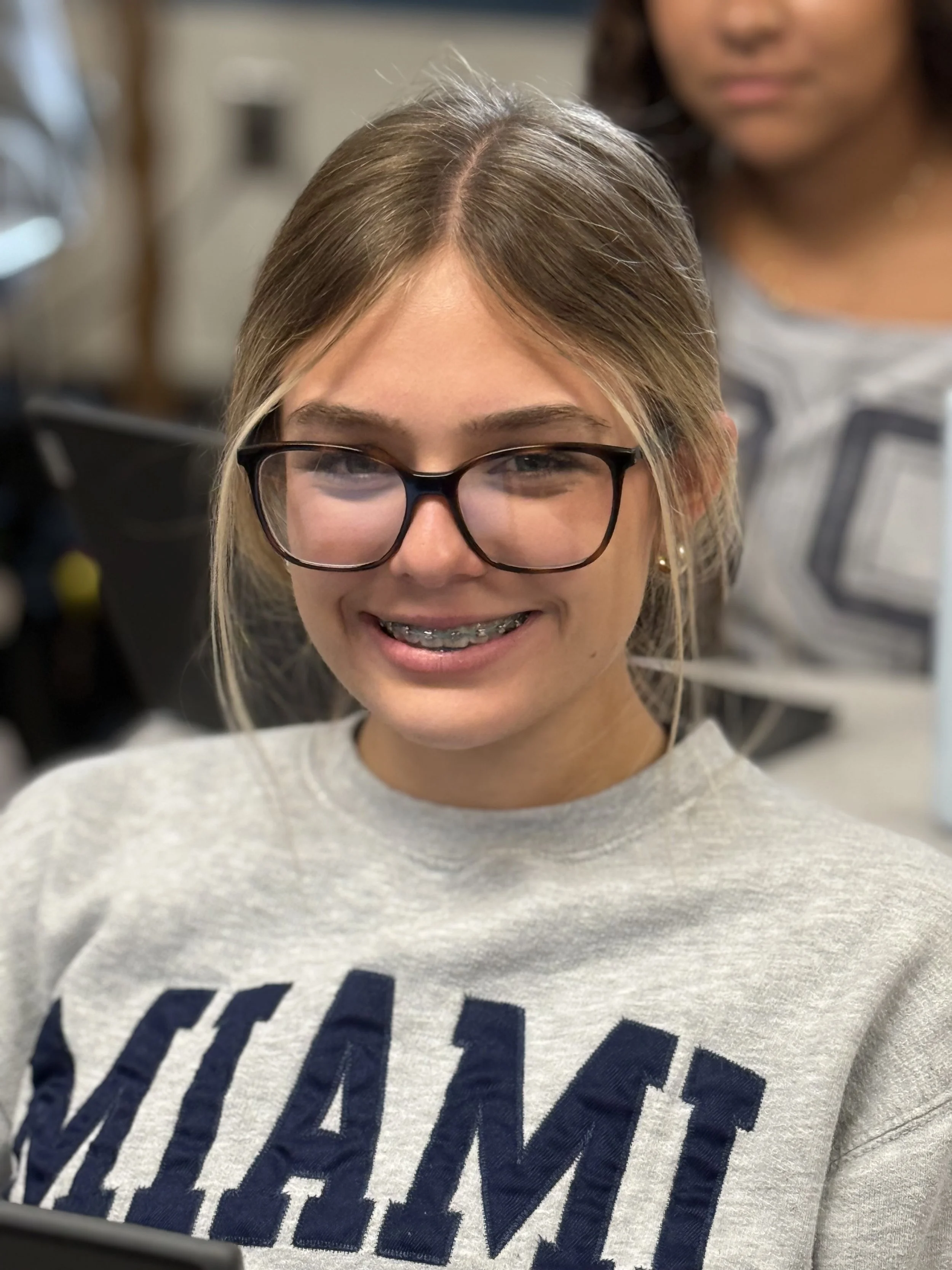 A young girl with glasses smiling showing braces, wearing a gray sweatshirt with dark blue text, and a woman is visible in the background.