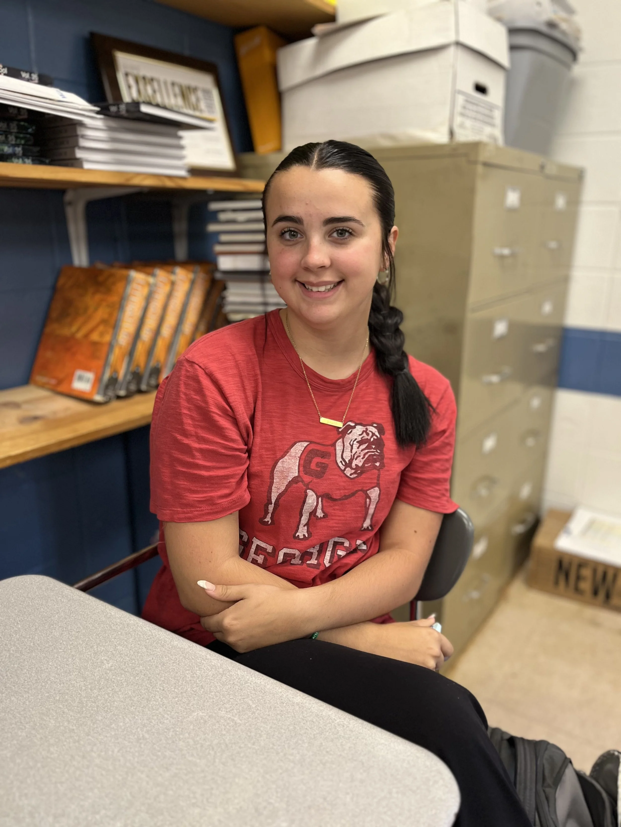 A smiling young woman with braided hair, wearing a blue t-shirt with a graphic and a necklace, sitting at a table in a well-lit room.